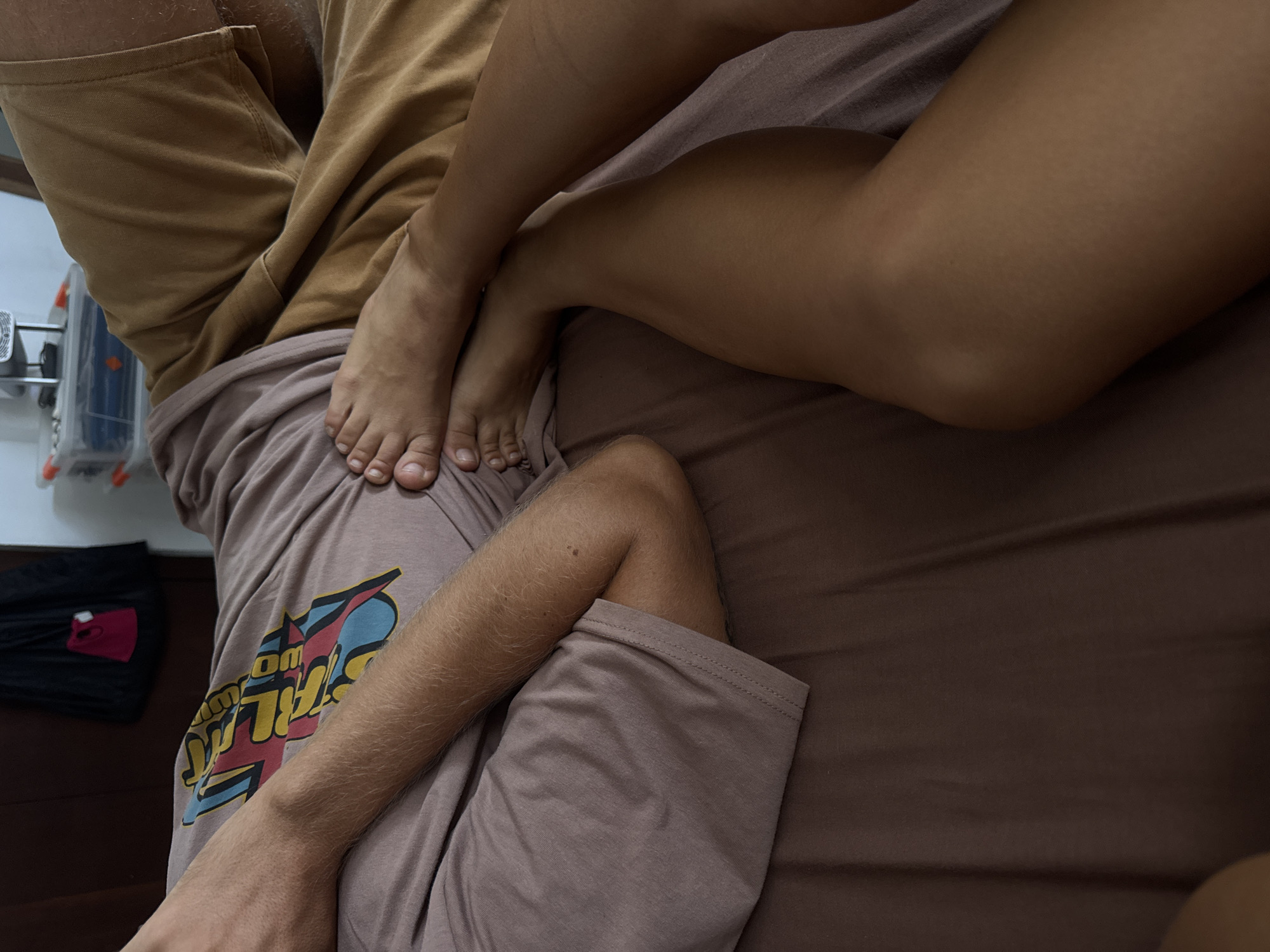 The image shows two individuals on a bed in what appears to be an indoor setting. One person is wearing a light brown t-shirt and darker brown shorts, with a graphic design on the shorts. The other person's leg is draped over the first person, revealing tan skin. The bed is covered with a brown sheet. In the background, there is a wall with an object attached, along with black clothing on the floor. The exact location is Goviyapana, Sri Lanka.
