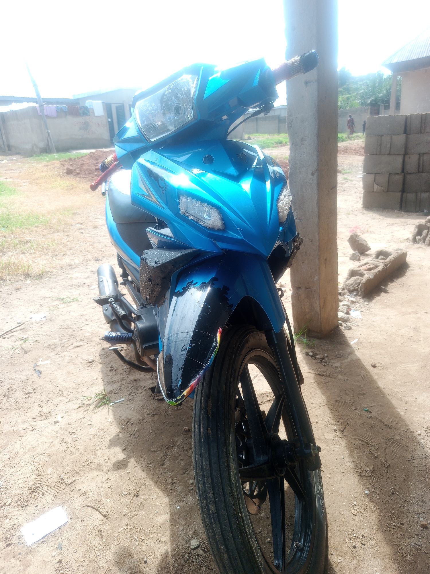 An image captured in the city of Keffi, Nigeria, displays a bright metallic blue underbone-style motorcycle parked on dry, light brown sandy earth. The motorcycle is positioned facing slightly towards the left of the frame, with its front wheel and fender prominently featured in the lower right quadrant. The front fender is black with a reflective rainbow-striped edge. The handlebars, seat, and exhaust pipe are partially visible. The front wheel features black alloy spokes and a ribbed tire.
In the immediate foreground, to the left of the motorcycle's front wheel, a small patch of sparse green grass and a rectangular white object are visible on the dusty ground. Long, distinct shadows are cast across the ground, originating from the motorcycle and a large, weathered concrete pillar positioned in the right mid-ground, partially obscuring the motorcycle's body.
Behind the concrete pillar, a stack of grey concrete building blocks is neatly arranged. Further back, scattered debris resembling broken concrete or mud bricks is present. In the background, two buildings are visible: one to the left with light-colored walls and a clothesline displaying laundry, and another to the right with a corrugated metal roof. Two human figures, appearing to be adults, are walking on a dirt path in the far background between the buildings. Distant trees and vegetation are visible behind the structures. The sky is overexposed and appears bright white, indicating strong sunlight conditions.