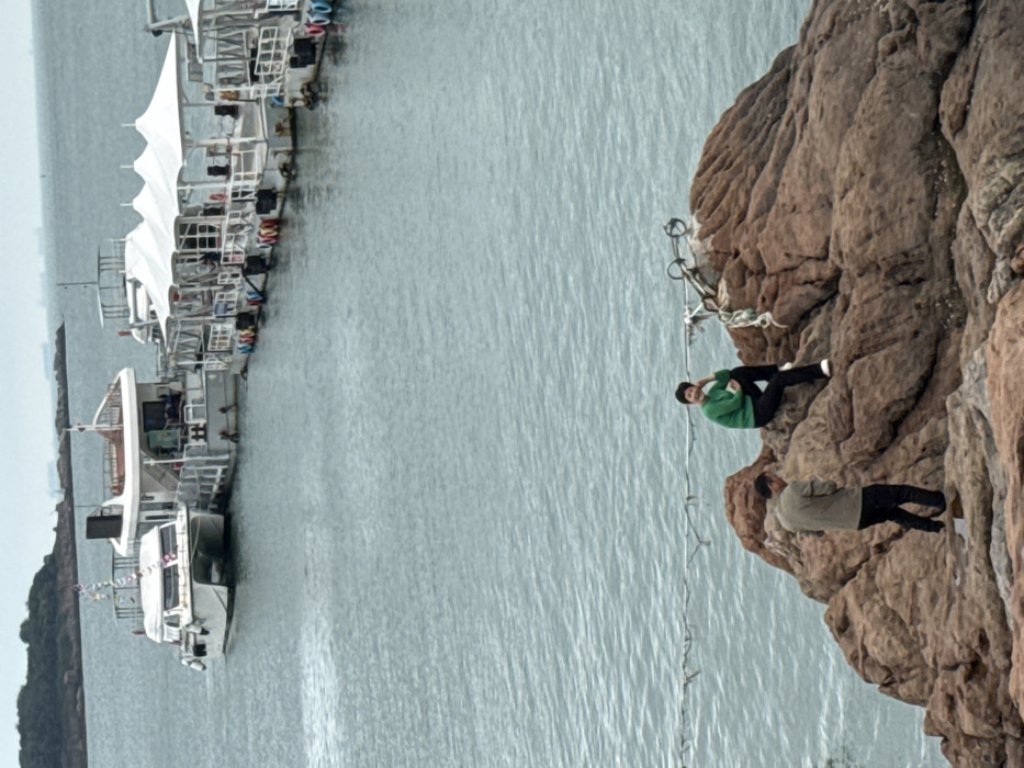 The image displays a coastal scene within Shinan District, China, under an overcast sky. In the foreground, a large, textured reddish-brown rock formation is present. Two individuals are on these rocks: a female in a green long-sleeved top and dark trousers is seated higher on the right, facing left with a slight smile; a male in a light brown jacket and dark trousers stands lower on the left, facing partially away from the viewer with his head angled downwards. A thin rope or cable extends horizontally across the midground, between the rocks and the water. In the background, calm grey-blue water extends to the horizon. Several boats are moored to a floating dock structure: a white catamaran-style yacht is partially visible on the left, and a series of at least four flat-decked recreational boats, each topped with white tent-like awnings, are arranged in a line to the right. Distant landmasses and faint vessel outlines are visible on the horizon.