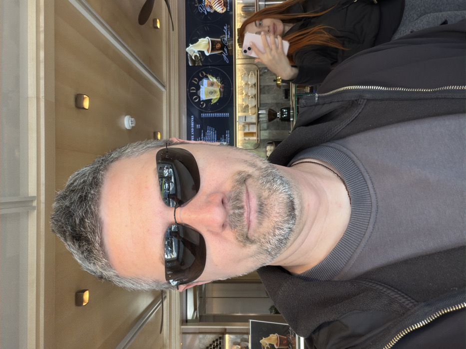 The image displays a selfie taken inside the "Goddo" cafe in Budva, Montenegro. In the foreground, a mid-adult male with short, spiky grey hair and a grey beard is present, wearing dark sunglasses, a grey crew-neck sweatshirt, and a black zipped jacket over it. A thin dark necklace is visible around his neck. The man is looking directly at the camera, and his sunglasses reflect an outdoor scene with vehicles.
In the background, behind a counter with various cafe equipment including a coffee grinder and stacked white cups, a female individual with long, reddish-brown hair is visible. She is wearing a dark jacket and grey trousers, holding a light pink smartphone with her right hand, angled as if capturing a mirror selfie. Her face is partially obscured by the phone.
The cafe environment features a wooden paneled ceiling with multiple recessed square light fixtures and a white smoke detector. A black ceiling fan is partially visible on the right side. On the wall behind the counter, a menu board titled "COFFEE AND DRINKS" prominently displays the "GODDO" brand logo, along with illustrated beverages and prices in Euros, such as "Hot Chocolate 2.50€."