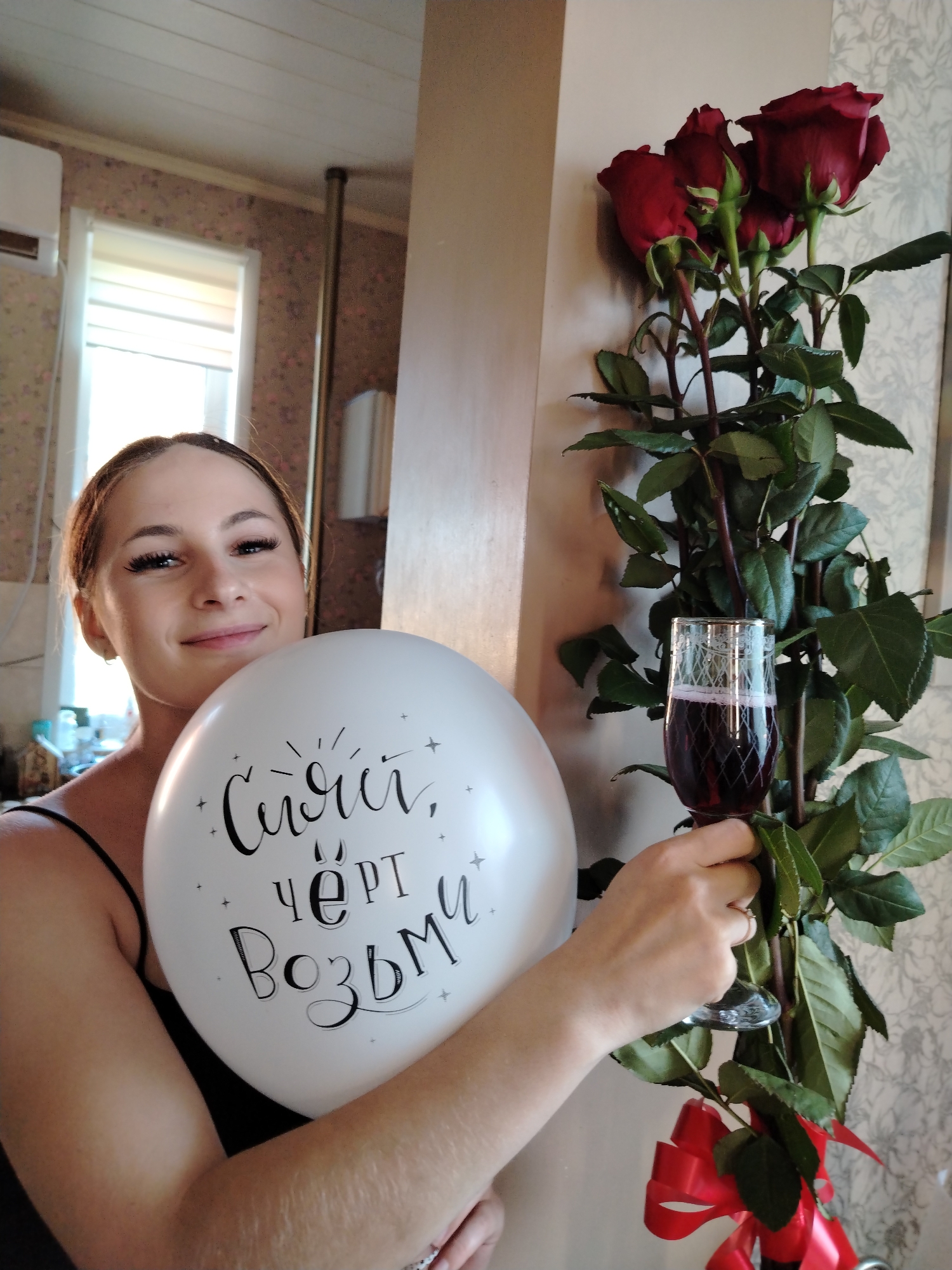 A woman stands indoors holding a white balloon with black lettering and a glass of red wine, next to a bouquet of red roses. The woman is wearing a black sleeveless top. The background shows a wall with patterned wallpaper, a window with blinds, and a ceiling with wooden planks. The scene appears to be a domestic setting.