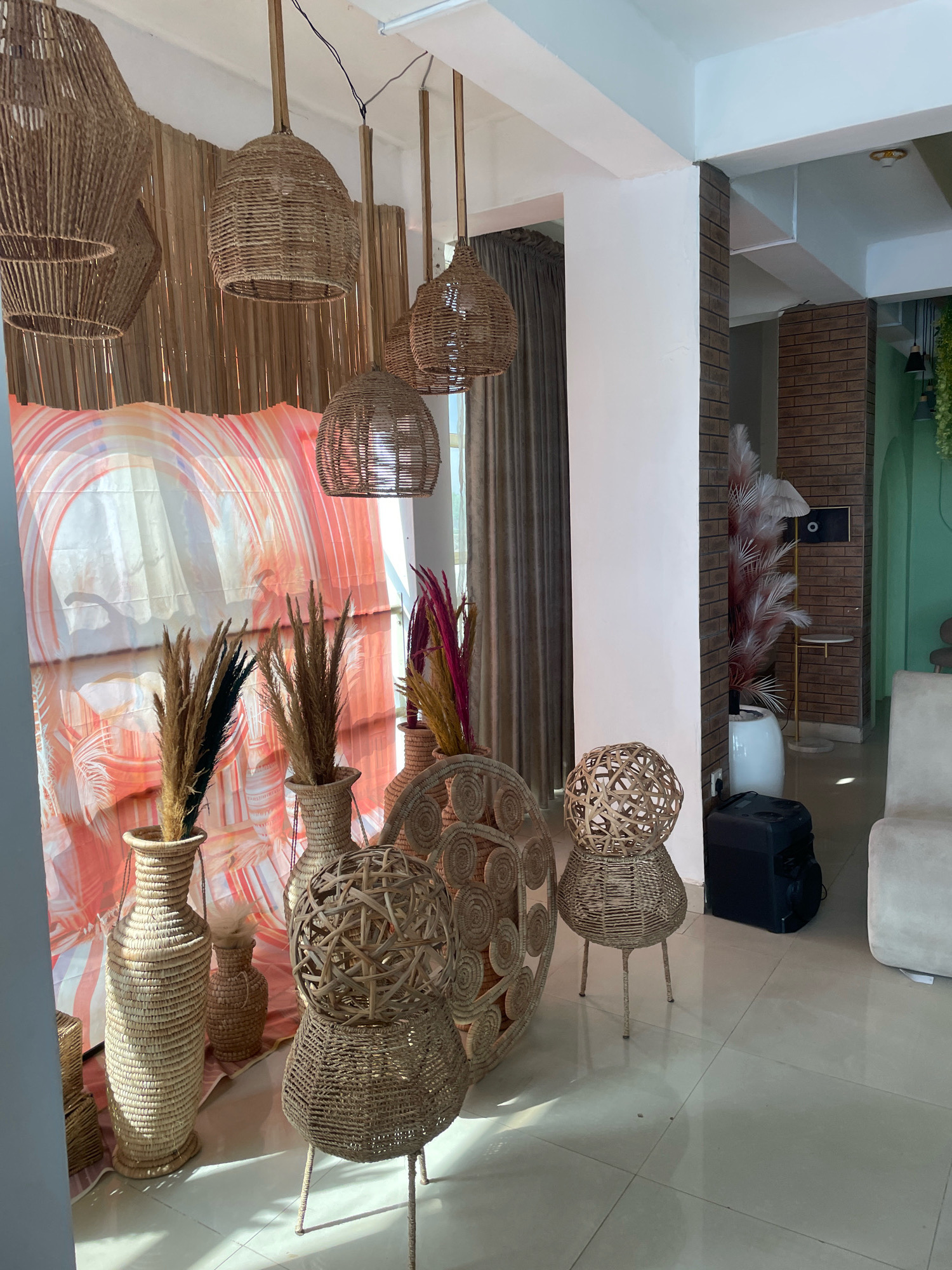 The image depicts an interior space, likely a shop or event venue, decorated in a bohemian style. The dominant materials are natural fibers like rattan and jute, used for furniture, lighting fixtures, and decorative elements.
The scene features several woven rattan pendant lights hanging from the ceiling. In the foreground, a large, tall woven vase holds dried reeds with dark and light brown tips. Next to it is a smaller woven vase also containing dried reeds. A large circular woven wall hanging with intricate patterns is visible behind these vases. To the right, a unique woven stool or side table is present, with a spherical top and a bulbous base supported by thin legs. Another similar but smaller woven spherical object sits atop a wider woven base in front of it. Further to the right, a plush cream-colored sofa is partially visible.
In the background, a vibrant, abstract textile with swaths of orange and pink creates a backdrop. A brick wall is visible, along with a mint green wall and a glimpse of greenery hanging from the ceiling, suggesting it might be a plant. A white cylindrical planter with wispy, pink and white decorative plumes stands near the brick wall, and a black speaker or equipment case is on the floor beside it.
The lighting suggests it is daytime, with sunlight casting shadows on the floor, indicating a bright but potentially diffused light source. The overall ambiance is warm, airy, and embraces a natural, rustic aesthetic. There is no visible text in the image. The location context is Ibadan, Nigeria.