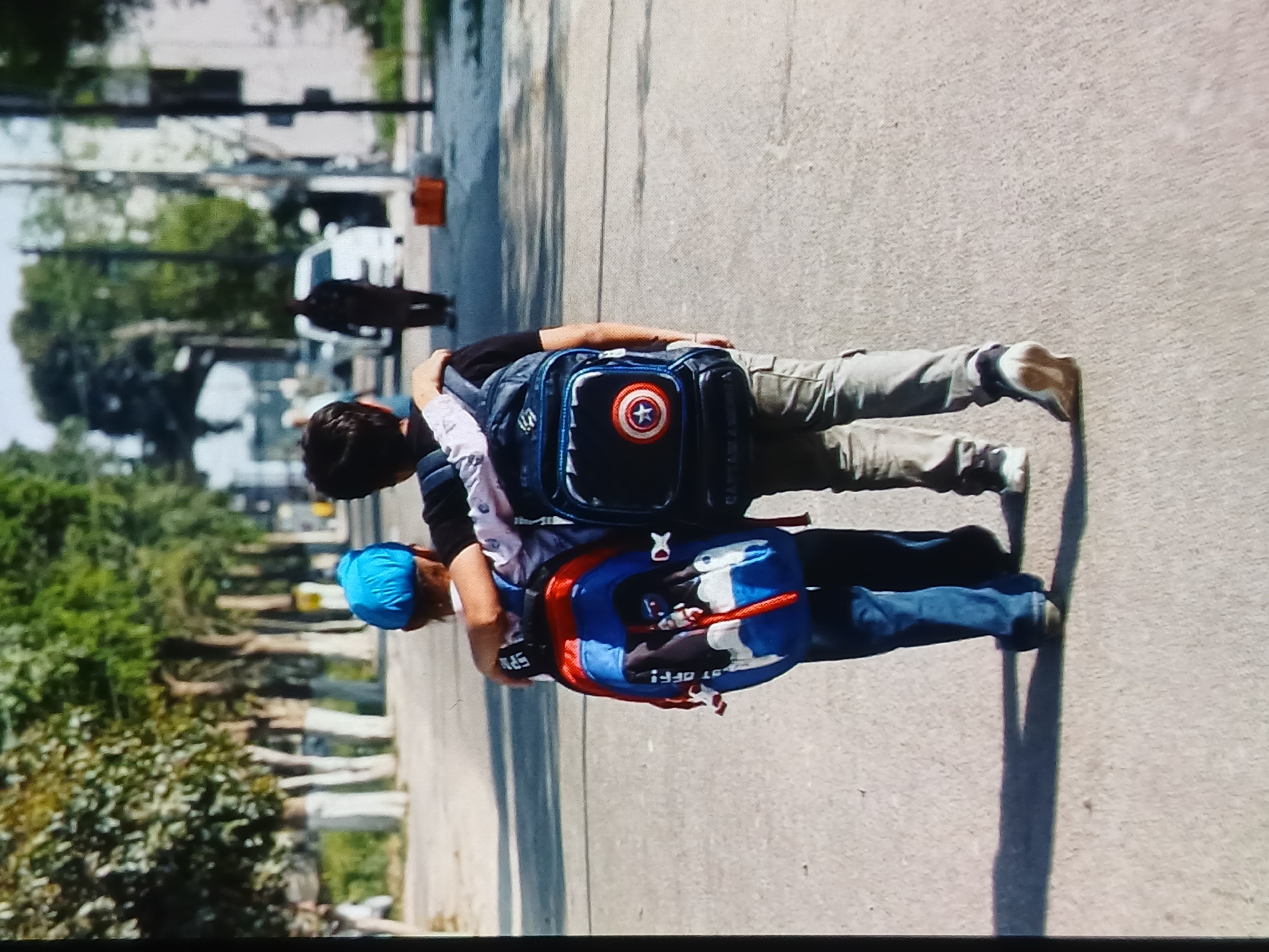 Two children, one wearing a blue headscarf, walk away from the camera with their arms around each other's shoulders. Both are carrying backpacks. The child on the left has a blue and red backpack with cartoon characters on it. The child on the right has a dark blue backpack with a Captain America emblem. They are walking on a paved road with trees and buildings in the background. The scene appears to be in a city environment. The image quality is low, with visible pixelation. The specific location in Setia Bakti, Indonesia, cannot be determined from the visual information provided.