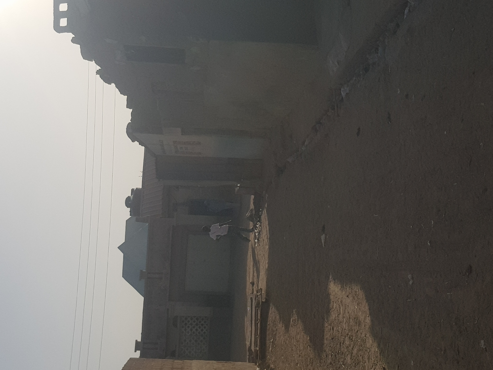 The image depicts a street scene in Dutsinma B, Nigeria. The dirt street runs along a row of buildings of varying heights and designs, constructed of aged materials. Two people are visible on the street. One person, likely a male, is wearing a light-colored shirt and dark pants, and appears to be standing next to a bicycle. The second person is standing close to the first person. Overhead power lines are visible in the sky. The image is taken in daylight, with a hazy or smoky atmosphere.
