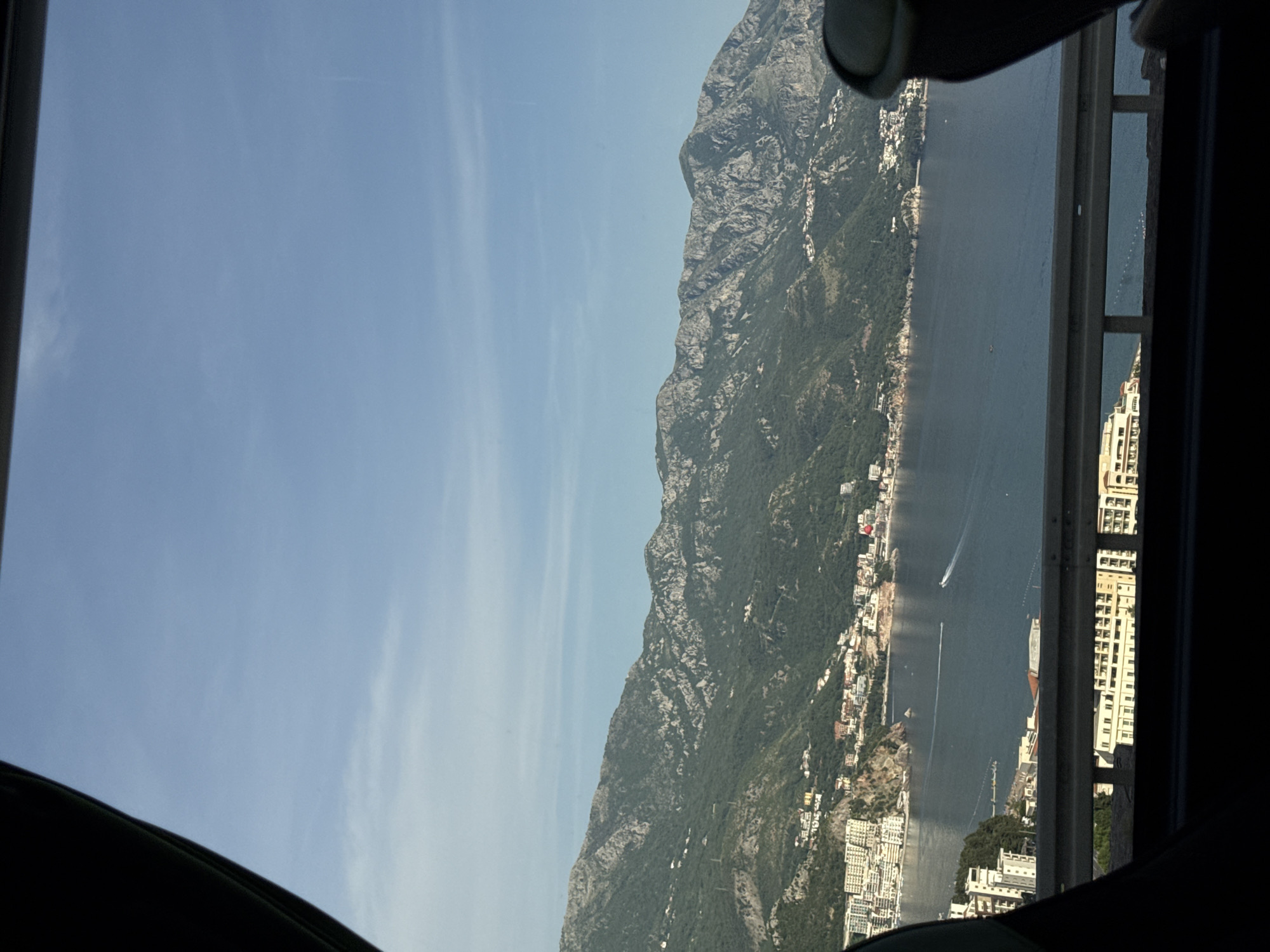 A vertical view from a car window captures the landscape of Boreti, Montenegro. The left of the frame showcases a clear blue sky with faint cloud wisps. Adjoining the sky is a rugged mountain range covered in greenery. Along the shoreline, buildings and structures form the town. The Adriatic Sea stretches beside the town, where a few boats dot the water. The frame is bounded by the edges of the car window.