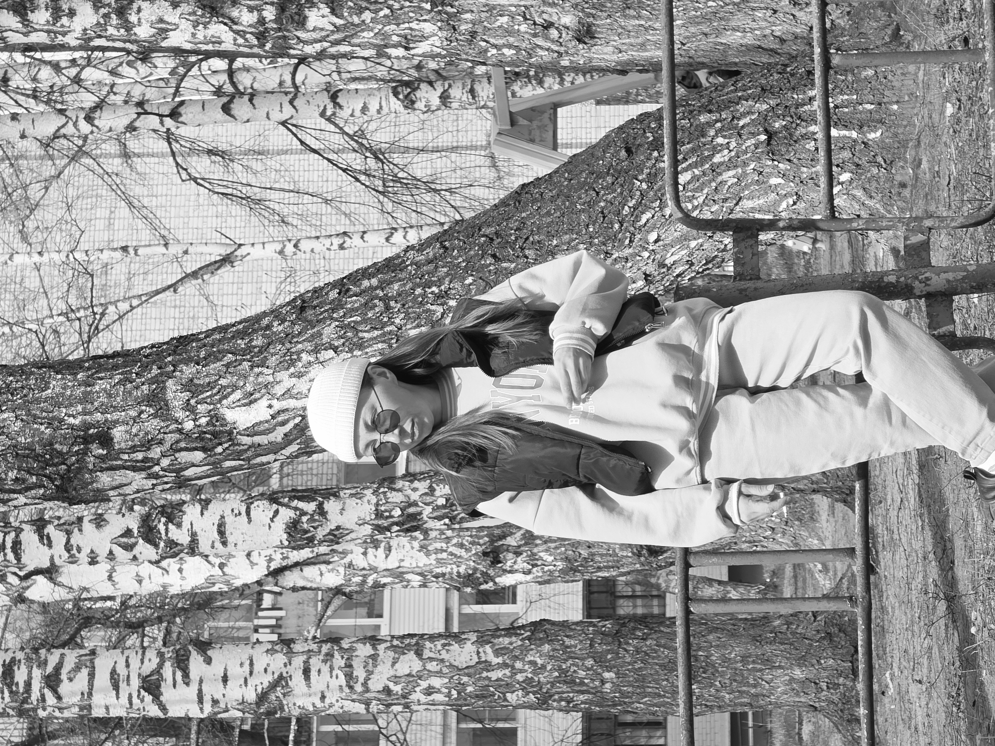 A black and white photograph, originally rotated, depicts a young person standing outdoors amidst trees, with residential buildings in the background. The person, likely female, is positioned towards the center-right of the frame, facing slightly left. She wears a light-colored hooded sweatshirt, light-colored pants, a dark vest or jacket, a white beanie, and round sunglasses. Her long hair is visible under the beanie. Her arms are bent, and she appears relaxed.
She stands in front of a tree with rough, dark bark, which dominates the right side of the image. To her left, extending into the mid-ground and background, are several distinctive birch trees with their characteristic white bark and dark markings. Their bare branches, along with the overall atmosphere, suggest late winter or early spring.
In the background, behind the trees, are multi-story apartment buildings with visible windows and brick patterns, indicating an urban or suburban setting. A metal structure, possibly a playground ladder or part of a fence, is partially visible to the right of the person, next to the dark-barked tree.
The bright lighting suggests it is daytime and sunny, casting subtle shadows. The overall scene conveys a casual outdoor moment in a park or residential area. On the person's hoodie, faint text "YO" and "ED G" can be seen. The image is set in Chaikovsky, Russia.