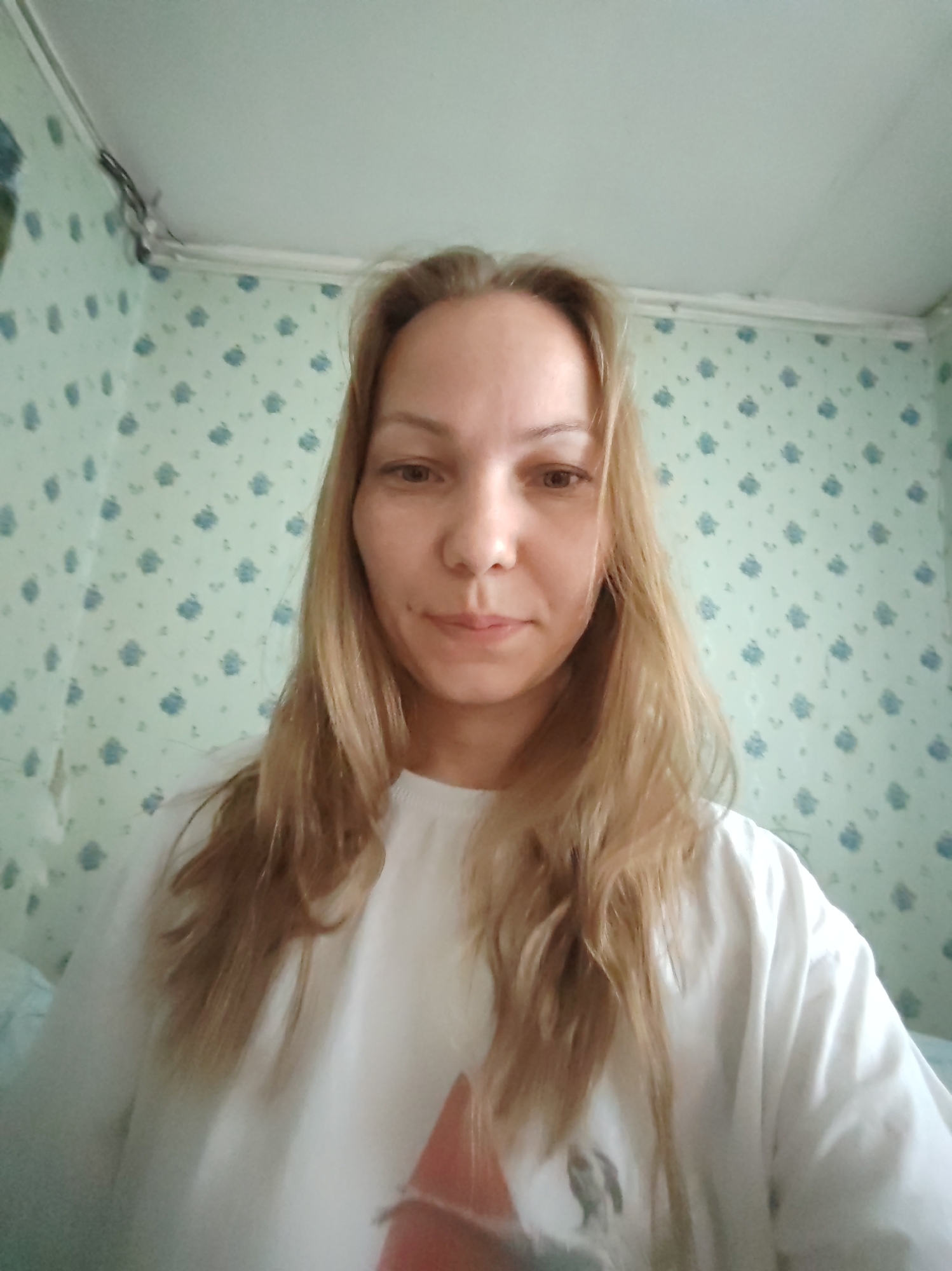 A head-and-shoulders, front-facing selfie of a woman with long, blonde hair. She is wearing a white t-shirt with a graphic design on it. The background is a light blue wallpaper with a repeating pattern of small, darker blue flowers. The scene appears to be indoors, possibly in a residential setting. The lighting is natural and appears to be coming from above. The overall impression is that of a casual, everyday moment. This image was taken in Chukhlem, Russia.