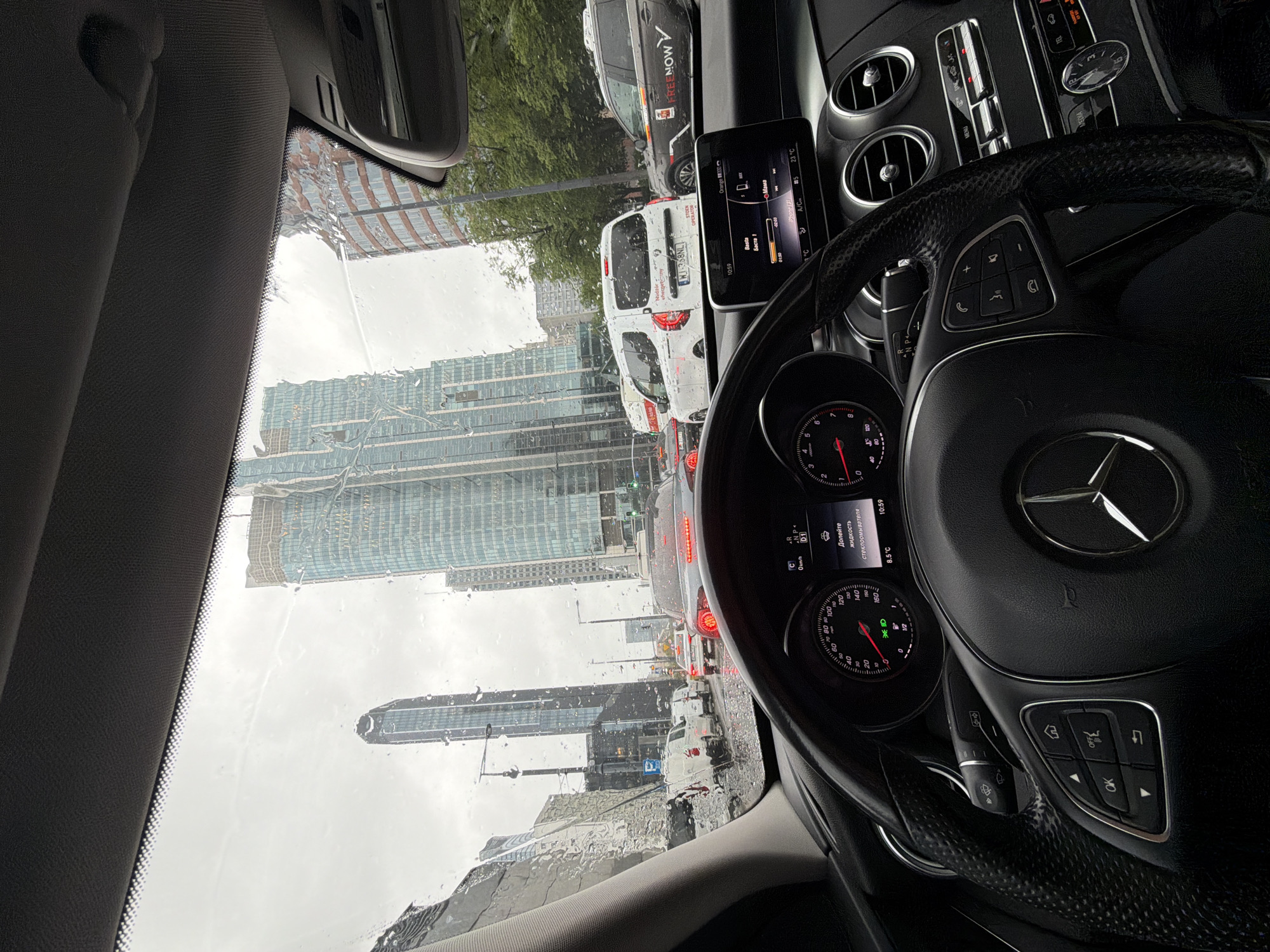 Captured from inside a car, the photograph portrays a rainy day in a city dominated by modern architecture. Towering skyscrapers, one with a distinctive elliptical apex, punctuate the skyline visible through the water-streaked windshield. The rain obscures a clear view of the city.
Within the car's interior, the focus is on the dashboard, with the Mercedes-Benz steering wheel prominently displayed. The instrument panel shows information like speed and temperature. The central console screen displays navigation or infotainment information. Traffic is visible outside the car, with other vehicles on the road. The overall impression is of urban congestion on a wet day.