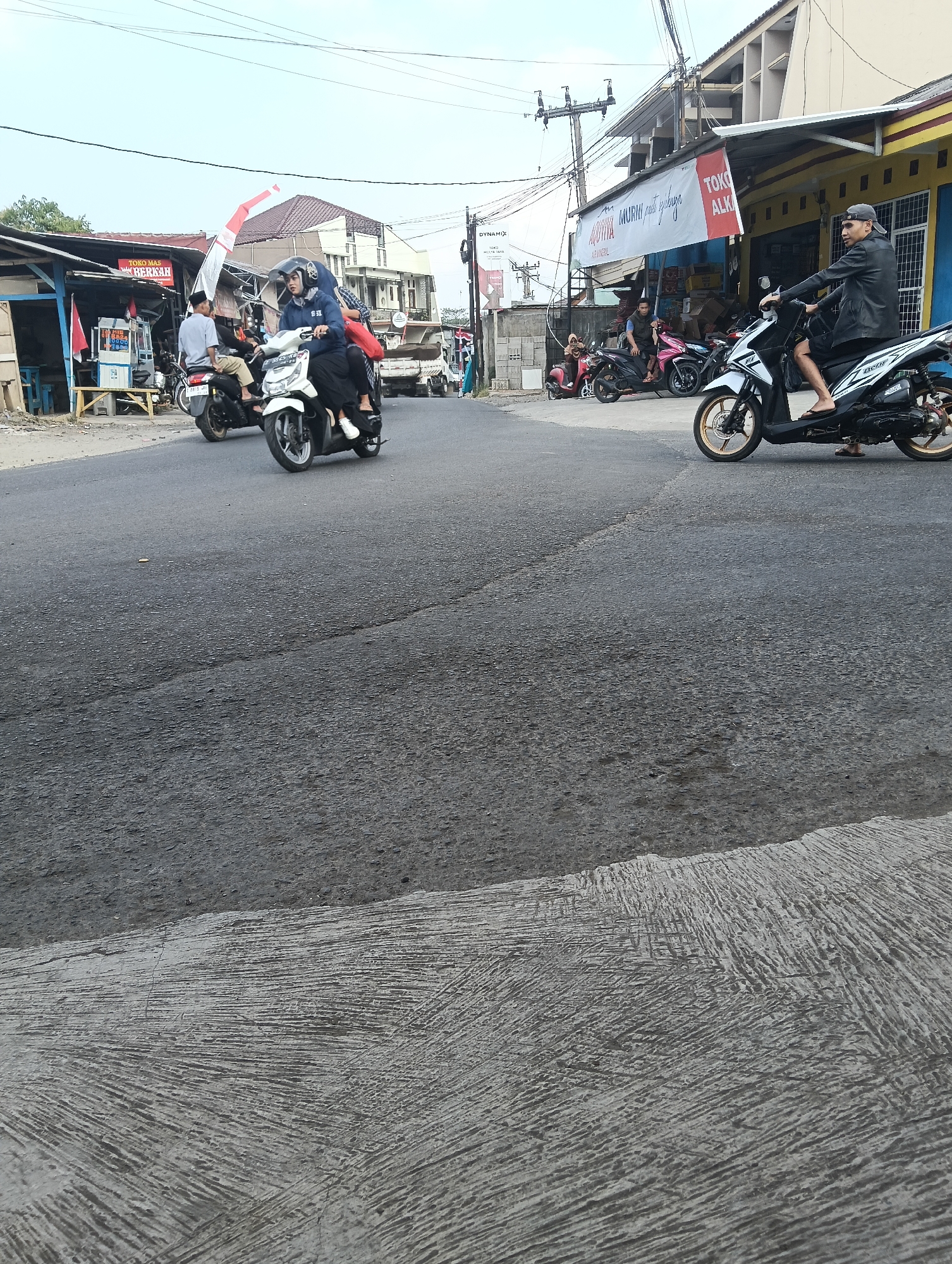 In Puspahiang, Indonesia, a street scene unfolds with multiple motorcycles and pedestrians navigating the asphalt road. In the foreground, a person rides a white scooter with gold rims, wearing a black jacket and a grey baseball cap, looking towards the right. Behind them, a pink and black motorcycle is parked, with a person wearing a grey t-shirt seated on it. Further back, a white scooter carries two individuals, the passenger wearing a blue headscarf and black clothing, while the rider wears a light-colored shirt and black pants. To the left, another motorcycle is occupied by a person in a light-colored shirt. Shops line the right side of the road, with signage including "TOKO MAS BERKAH" and "TOKO ALKA." A truck is visible in the background. The sky is overcast, and power lines crisscross overhead. The road surface shows some cracks and texture.
