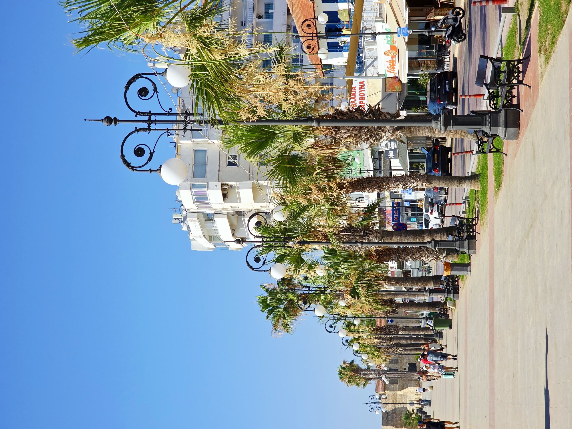The image depicts a sunny day along Finikoudes Beach promenade in Larnaca, Cyprus. A wide, paved walkway, bordered by grass and benches, runs alongside a row of palm trees. Beyond the trees, a street is lined with buildings, including shops and cafes. Several people are walking along the promenade, some appearing to be tourists. Cars and a scooter are visible on the street. The sky is clear blue.