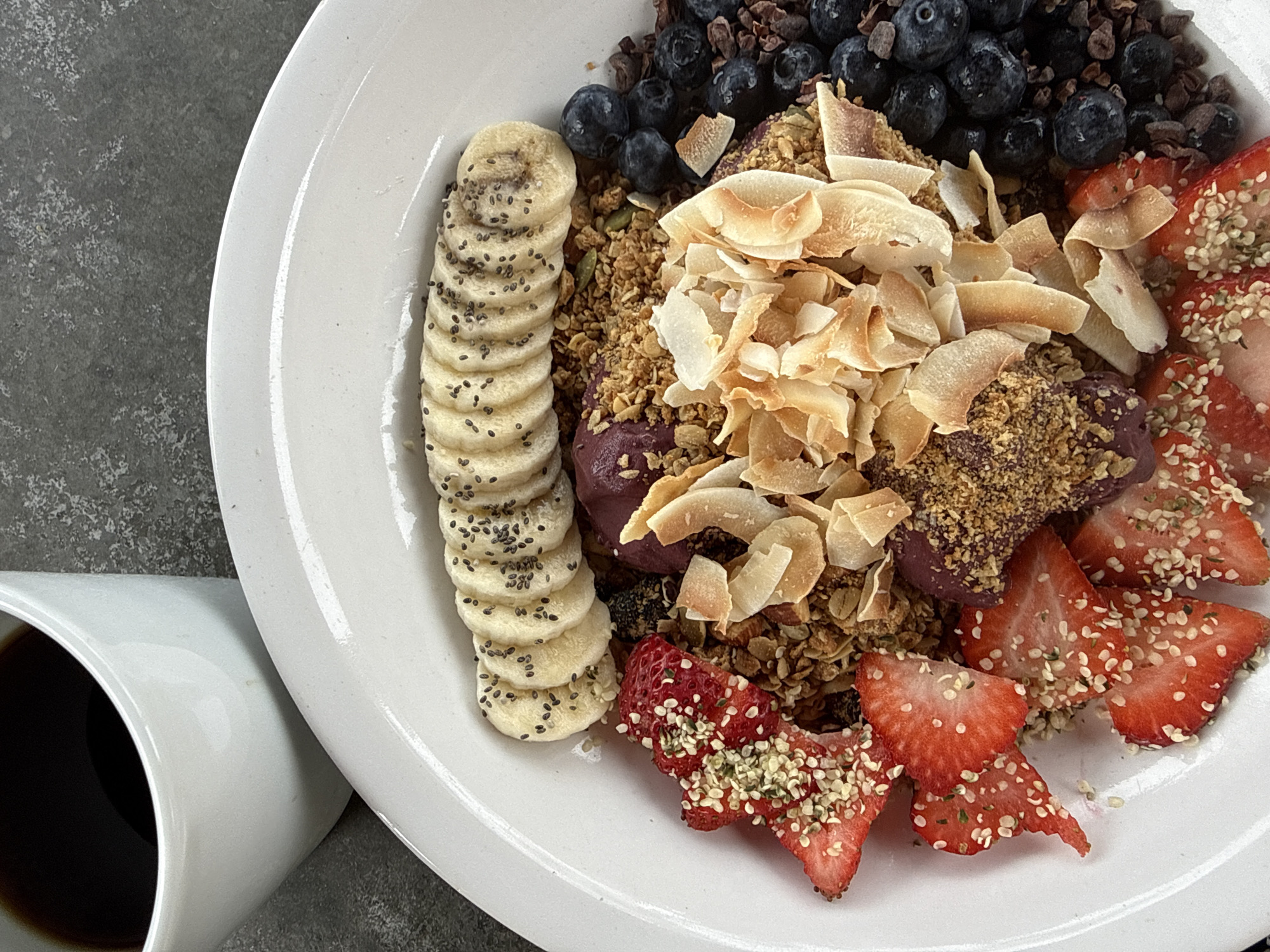 The image displays a healthy breakfast or brunch meal featuring an acai bowl and a cup of coffee.
**Subjects:** The main subject is a vibrant acai bowl filled with various colorful toppings, presented on a round, white ceramic plate. Alongside the plate, a simple white ceramic mug holds a dark liquid, which appears to be coffee.
**Activity or event taking place:** The image captures a moment of a prepared meal, ready to be consumed, suggesting a leisurely breakfast or brunch.
**Notable details:**
* **Acai Bowl Contents:** The acai base is a deep purple color, mostly covered by an abundant arrangement of toppings.
* Along the left edge, a neat line of sliced bananas is visible, each piece sprinkled with small black chia seeds.
* The upper right section features a generous portion of fresh blueberries, intermingled with dark, irregularly shaped pieces that resemble cacao nibs or dark chocolate.
* A significant amount of golden-brown granola, containing visible pumpkin seeds, forms a base for other toppings.
* Piled prominently in the center are light brown, flaky toasted coconut chips, adding texture and warmth.
* The lower right portion of the bowl is adorned with bright red, sliced strawberries, heavily sprinkled with light green or beige hemp seeds.
* **Coffee:** The white mug contains a dark, presumably black, coffee.
* **Setting:** The plate and mug rest on a dark gray, subtly textured surface, likely a stone countertop or table, which provides a neutral and modern backdrop.
* **Presentation:** The food is artfully arranged, emphasizing its freshness and visual appeal. The meal appears wholesome and nutritious.
* **Time of Day:** The nature of the meal suggests it is consumed in the morning or early afternoon.
**Visible text:** There is no visible text in the image.