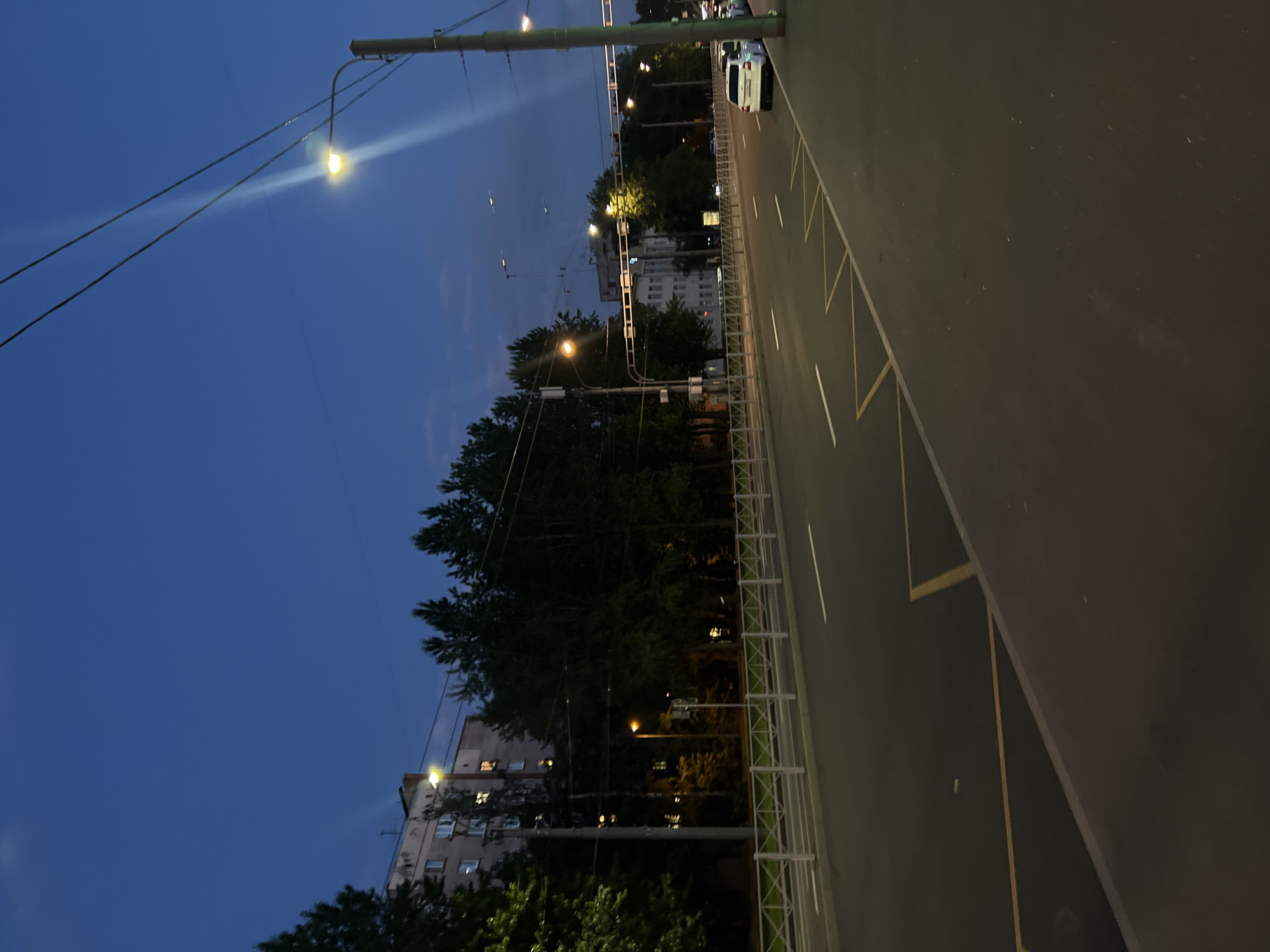 The scene depicts a city street at twilight or early night in the city of Unknown, Unknown. The sky above is a deep, uniform dark blue, with faint lighter patches and minimal cloud cover visible in the upper right.
In the mid-ground and background on the left, a multi-story, light-colored residential building (likely an apartment block) with multiple rectangular windows is visible. Several of these windows emit a warm yellow light, indicating interior illumination. Dark green coniferous and deciduous trees heavily line the left side of the street, partially obscuring the lower floors of the building.
The asphalt road, which appears dry, runs from the foreground towards the background, featuring at least two visible lanes. It is marked with white dashed lane lines separating traffic directions. Along the right-hand curb in the foreground and mid-ground, a series of yellow triangular markings (commonly known as "shark's teeth" or "give way" markings) are painted on the road surface, adjacent to a solid white line, suggesting a specific zone such as a bus stop or restricted parking area.
A white metal pedestrian barrier, composed of horizontal and diagonal bars, runs along the curb on the left side of the road, separating the roadway from a sidewalk and a line of trees. Similar barriers are visible further down the street on both sides.
Numerous utility poles and streetlights line both sides of the road. The streetlights emit a bright, warm yellow-orange light, creating visible light streaks in the air, especially prominent from the streetlight in the upper right quadrant of the image. Overhead electrical wires crisscross the sky, connecting to these poles. Traffic light gantries with overhead lights are visible in the mid-distance, spanning the road.
On the far right, a wide concrete sidewalk borders the road, and a single white sedan is parked parallel to the curb. Further down the street, additional vehicle lights and building structures are faintly visible through the trees and ambient darkness. No people are visible in the image.