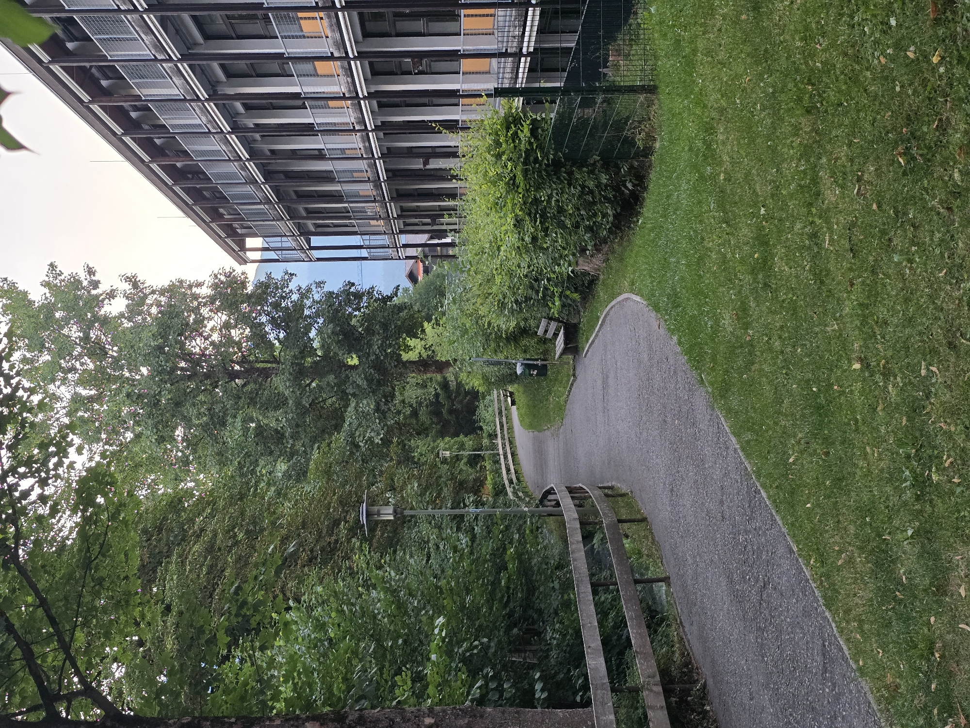The image depicts an outdoor scene in Garmisch-Partenkirchen, Germany. A paved pathway curves through a green, grassy area on the right, bordered by a dense hedge. To the left of the pathway, a wooden fence runs along the edge of a wooded area with lush green trees. A street lamp is visible along the pathway. In the background, a modern, multi-story building with balconies and numerous windows is situated. The sky is overcast, suggesting a cloudy day. No people are present in the frame.