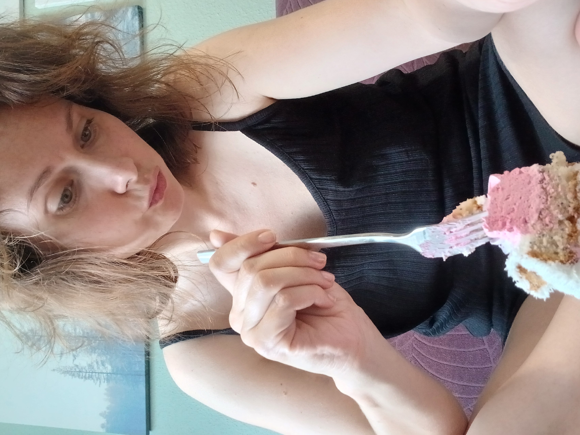 The image shows a woman sitting indoors, holding a fork with a piece of cake. The woman has brown hair and is wearing a black tank top. She is looking down at the cake. The background includes a painting on the wall and a purple cushion. The scene appears to be a domestic setting, possibly a living room or bedroom. There is no indication that this image is related to Yekaterinburg, Russia.