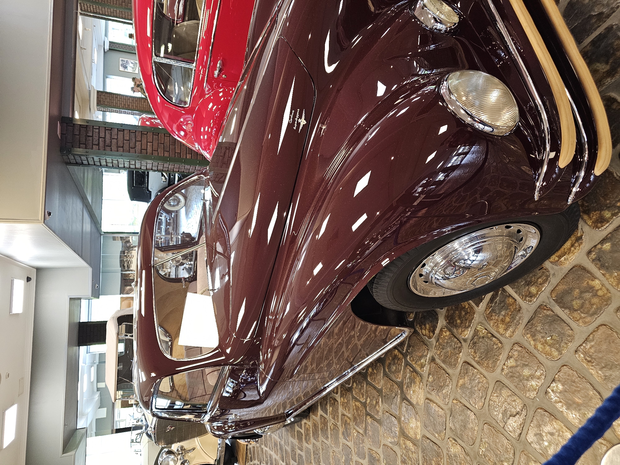 The image shows a close-up of two vintage cars in a museum setting. The primary car, in the foreground, is a maroon color with a highly polished finish, reflecting light and highlights. Its headlights are prominent, and a strip of wooden trim is visible along the front. The wheel features a chrome hubcap. The second car, in the background, is red. Both cars are parked on a cobblestone-like surface. A glimpse of the museum's interior is visible, with windows and architectural details suggesting the location might be in Архангельское, Russia, as museums are common in such historical places. The image is taken at a slight angle, giving a dynamic perspective of the cars.