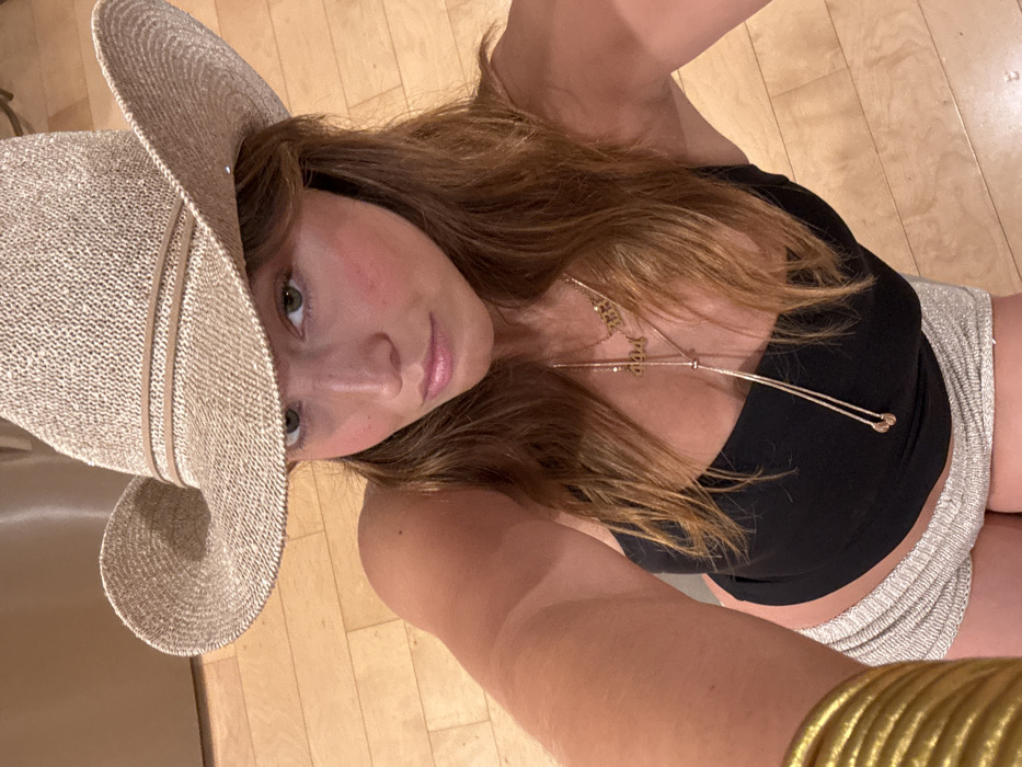 A woman with light brown hair and a tan complexion wears a large straw hat, a black top, and silvery shorts. She is adorned with gold necklaces and a golden bracelet. The background appears to be a wooden floor and part of a light-colored wall. The photograph is taken from a high angle, slightly tilted. The woman is looking towards the camera.