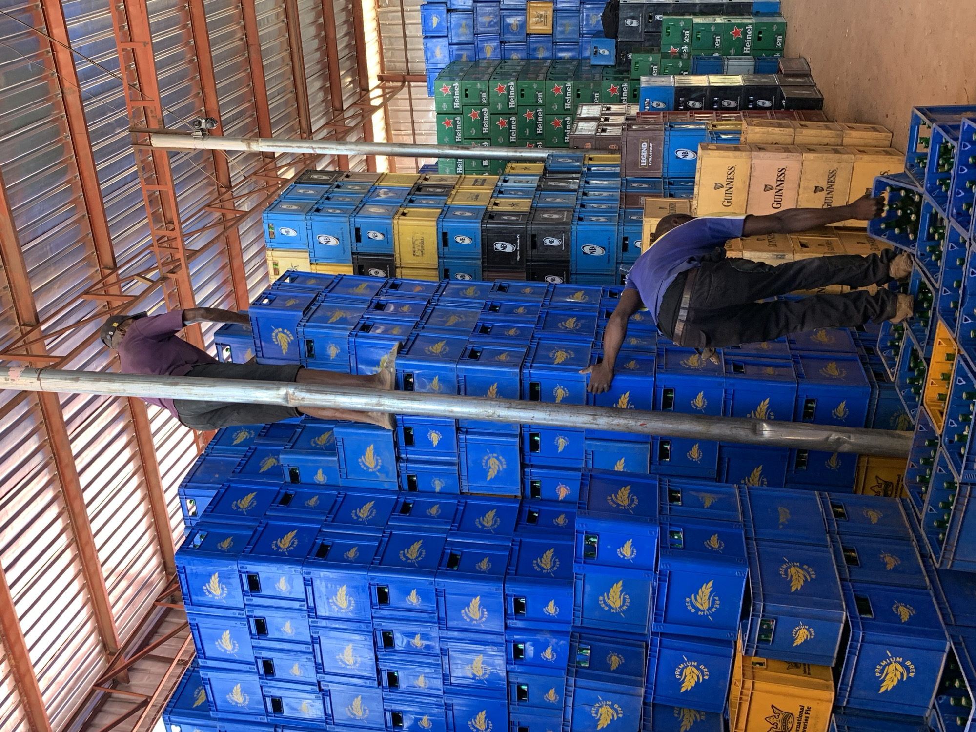 Two individuals are seen amidst stacks of beverage crates in what appears to be a warehouse-like structure, possibly located in Gura Top, Nigeria. The crates are various colors (blue, yellow, green, black), with labels such as "GUINNESS," "Heineken," "LEGEND," and "PREMIUM BREW," suggesting they contain alcoholic beverages. The individuals are reaching across or working with the crates. The building has an orange metal roof structure.