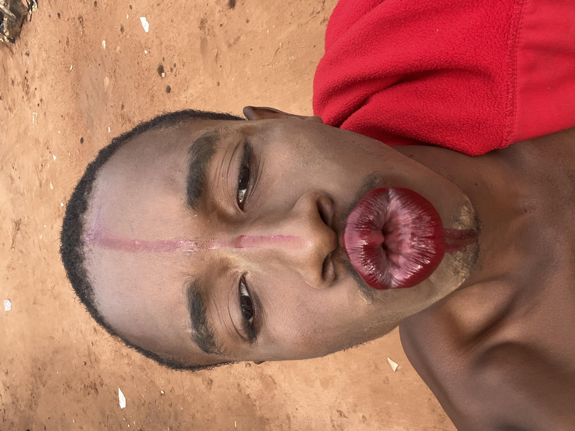 A close-up portrait shows a young Black male against a sandy or dirt ground background, contextualized in Onitsha, Nigeria. The subject looks directly at the camera with a neutral expression. His most prominent feature is a large, shiny, deep red, puckered object affixed to his lips, making them appear greatly exaggerated and distorted. A reddish-pink line is painted horizontally across his forehead and down the bridge of his nose. Faint lighter-colored material, possibly makeup or paint, is visible on parts of his face.
The man has short, dark hair. A bright red, textured fabric, likely a blanket or towel, is visible on the right side of the frame, near his shoulder and neck. The background consists of light brown or reddish-brown dirt with scattered small debris, indicating an outdoor, unpaved setting. The lighting appears to be natural daylight. There is no visible text. The image captures a posed moment, possibly for artistic expression or a cultural event.