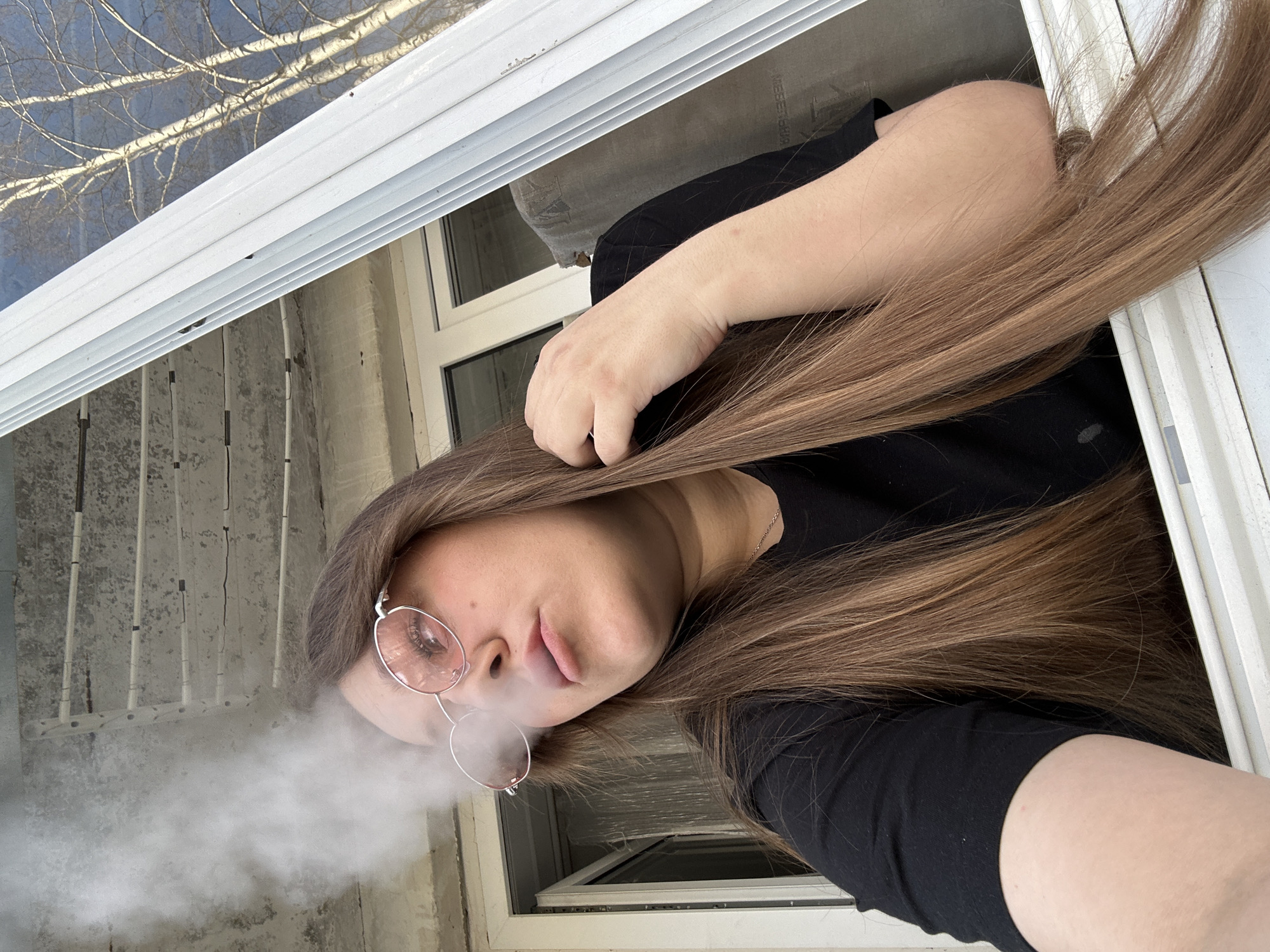 A woman with long brown hair and round glasses is exhaling smoke while leaning out of a window. She is wearing a black shirt. The window frame is white. Outside the window, there's a view of bare tree branches and the side of a light-colored building. The location is Chaikovsky, Russia.