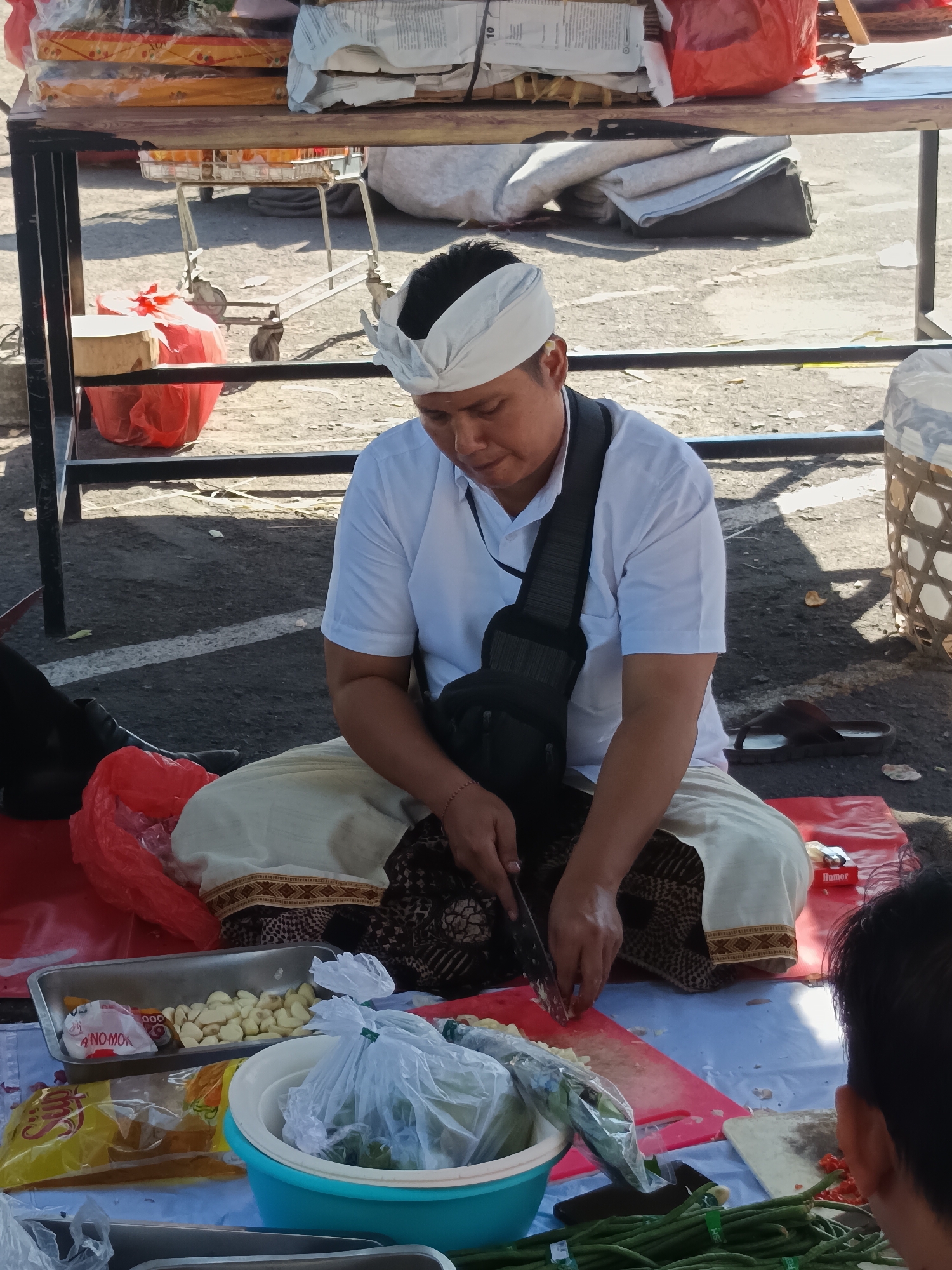 In Denpasar, Indonesia, a man wearing a white shirt and a white headcloth sits cross-legged on the ground, preparing food at an outdoor market stall. He is using a knife to chop ingredients on a red cutting board. In front of him are various food items, including a tray of garlic, a blue bowl with plastic-wrapped vegetables, and several packages of what appear to be spices or dry goods. A red plastic bag is also visible. The man has a black bag slung across his chest. Behind him, a wooden table holds stacks of packaged goods, and to the right, a woven basket is partially visible. The ground is paved and appears to be asphalt. The scene suggests a bustling market environment with a focus on fresh produce and ingredients.