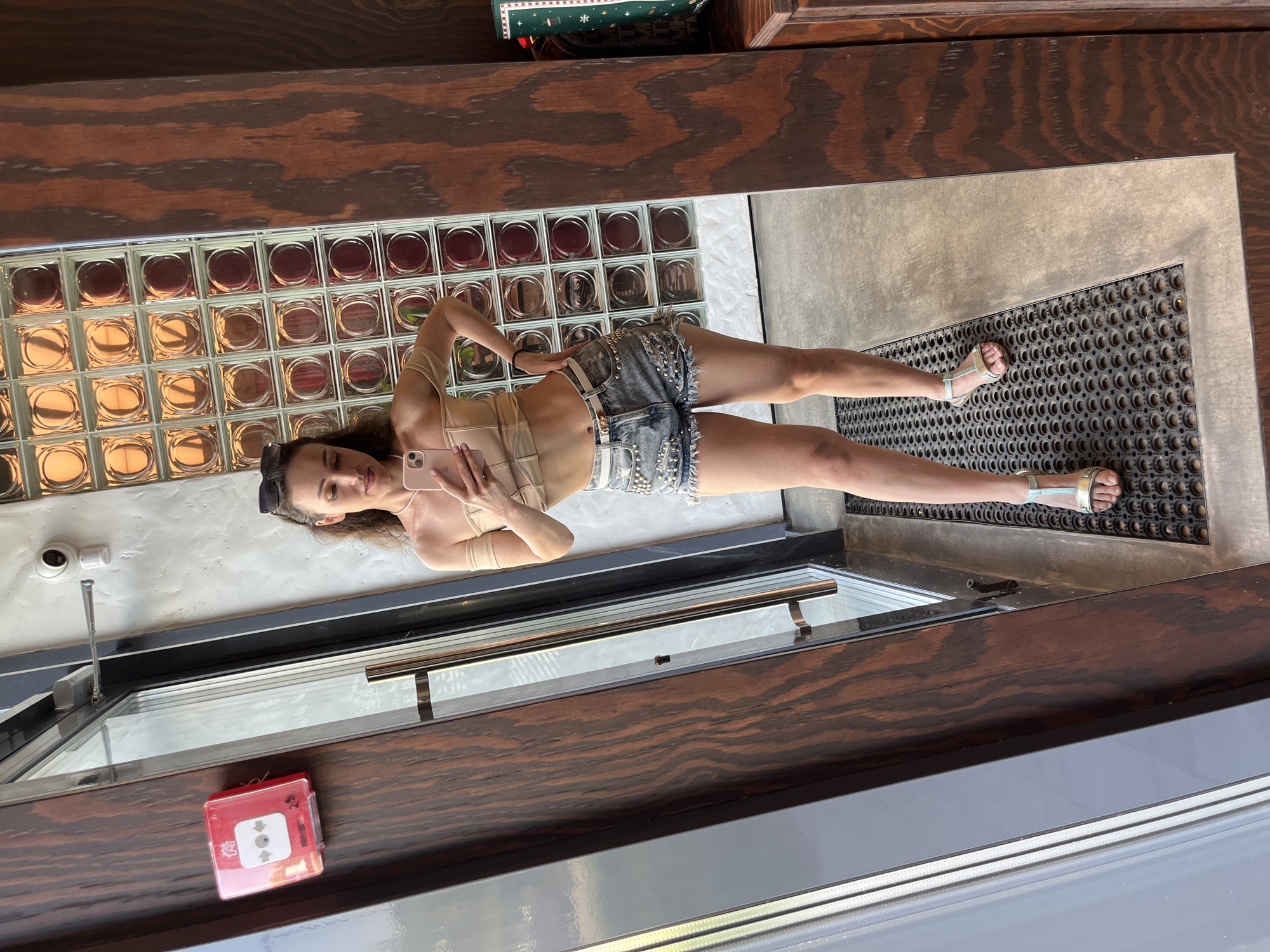 In Kazan, Russia, a woman stands in front of a mirrored surface, taking a selfie. She is wearing a light-colored, off-the-shoulder top and distressed denim shorts adorned with silver studs and a white belt. Her hair is long and wavy, and she is wearing sunglasses pushed back on her head. The background consists of a wooden door frame, a wall with glass blocks lit from behind, and a security camera mounted on the wall. A red fire alarm box is visible on the door. The floor is covered with a dark, circular mat.
