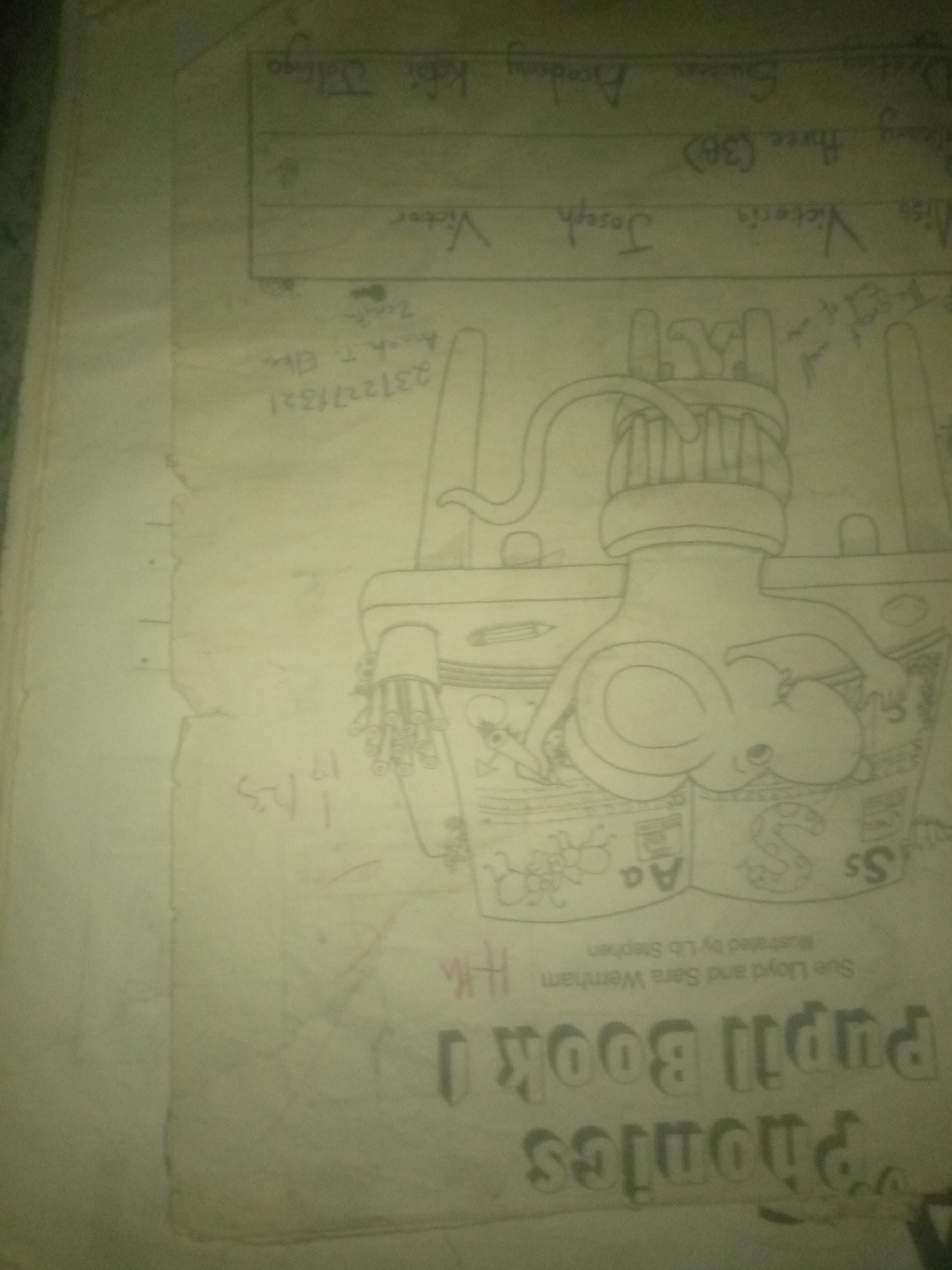 The image displays a worn, hand-drawn cover of a "Phonics Pupil Book 1." The cover is predominantly white with visible creases and smudges. The title is written in large, stylized capital letters in a dark color, appearing as "Pupil BOOK 1 Phonics." Below the title, smaller text reads "Sue Lloyd and Sara Wernham Illustrated by Liz Stephen." The central image is a detailed line drawing of what appears to be a stylized head or mask with intricate details, including what might be eyes, a mouth, and a headdress or hair-like elements. The drawing is in black pencil or ink. To the left of the drawing, there are handwritten names and numbers, including "2312277321," "Amak T. Ellis," and "Victor." Higher up on the page, there are more handwritten words, possibly names or class entries, such as "Victor Joseph," "Miss Victoria (38)," and "Mrs. Guster Academy." The handwriting suggests a school or educational context. The image does not provide any visual cues or contextual information to determine its location in Jauro Yinu, Nigeria, or any specific urban environment.