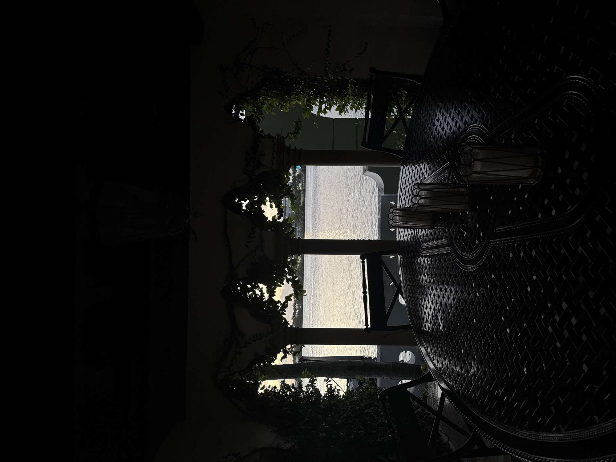 The image, taken at night in Miami Beach, United States of America, shows a partially illuminated outdoor patio. An intricately designed dark metal table dominates the right side of the frame, with a few chairs surrounding it. Through a series of three arched openings framed by pillars, a body of water reflecting light can be seen. Vines and foliage partially obscure the pillars and openings. The left side of the image is predominantly dark, suggesting the interior of the building or a covered area.