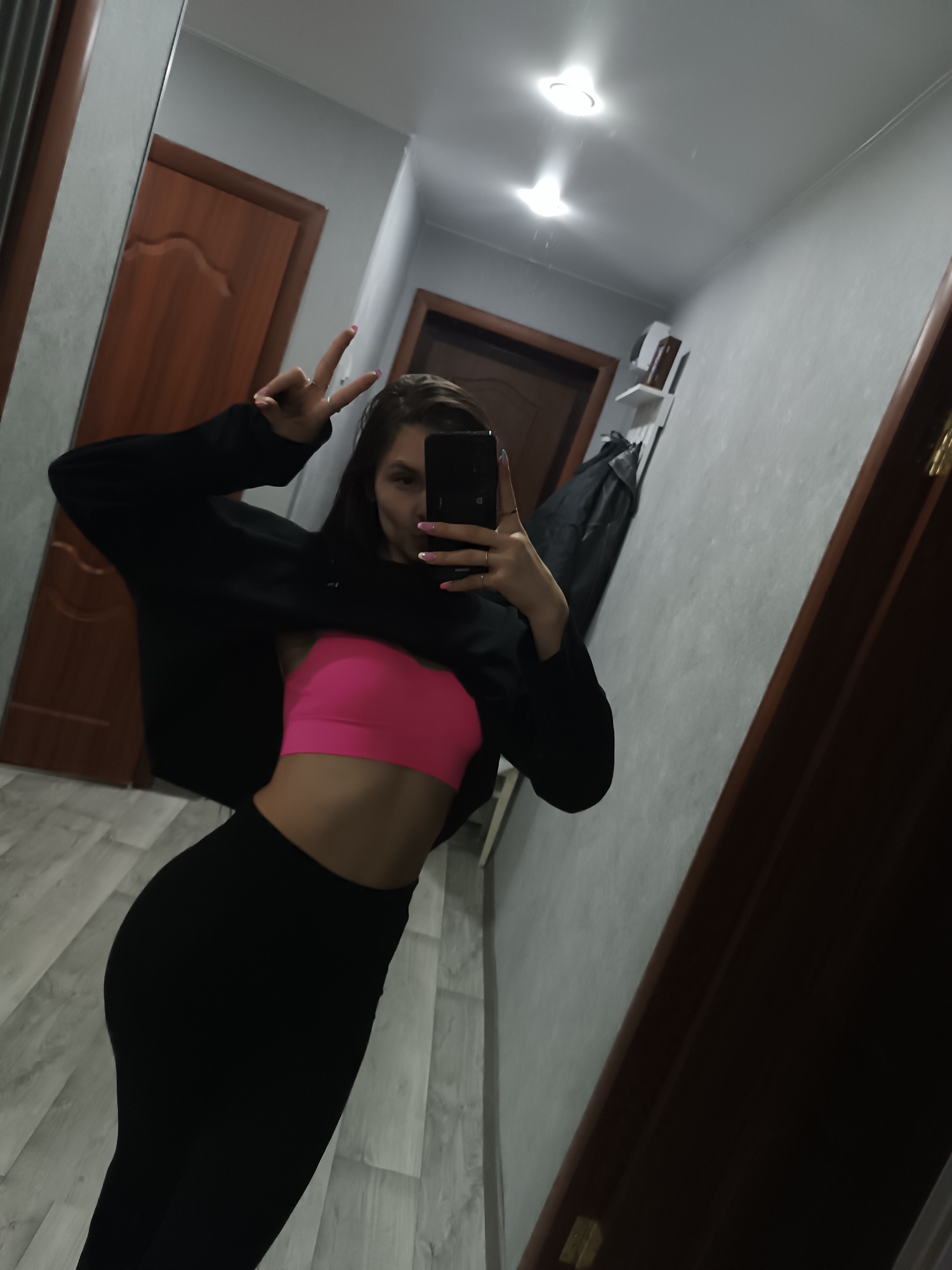 A woman is taking a selfie in a hallway. She is wearing a pink sports bra and black leggings, and a black sweater is draped off her shoulders. Her left hand is raised, making a peace sign. The hallway is narrow with light gray walls. There are two closed wooden doors visible. A light fixture is visible on the ceiling. A garment is hanging on a rack in the background. The floor is light-colored. The exact location within Chaikovsky, Russia, is not discernible from the image.