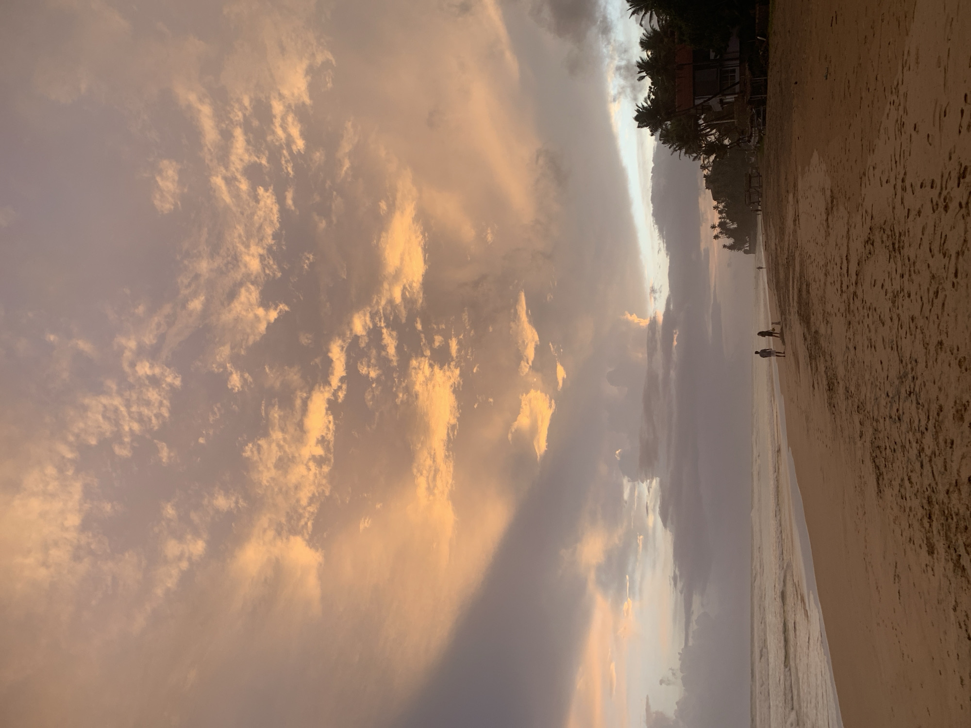 The image depicts a serene beach scene at dusk in Kathaluwa, Sri Lanka. The sky is filled with dramatic clouds painted in hues of orange and pink, with softer grey clouds interspersed. The ocean is calm, with gentle waves rolling onto the sandy shore.
In the foreground, the beach is wide and sandy, with footprints scattered across its surface, indicating recent human activity. Two figures, appearing to be a couple, are walking along the water's edge, silhouetted against the soft light. A small dog walks beside them. Further down the beach, another solitary figure is visible walking.
To the right of the frame, a row of palm trees lines the edge of the beach, with a building complex nestled among them. The building appears to be a resort or guesthouse, with a reddish-tiled roof and some thatched structures visible. The overall atmosphere is peaceful and tranquil, capturing the beauty of a tropical sunset.