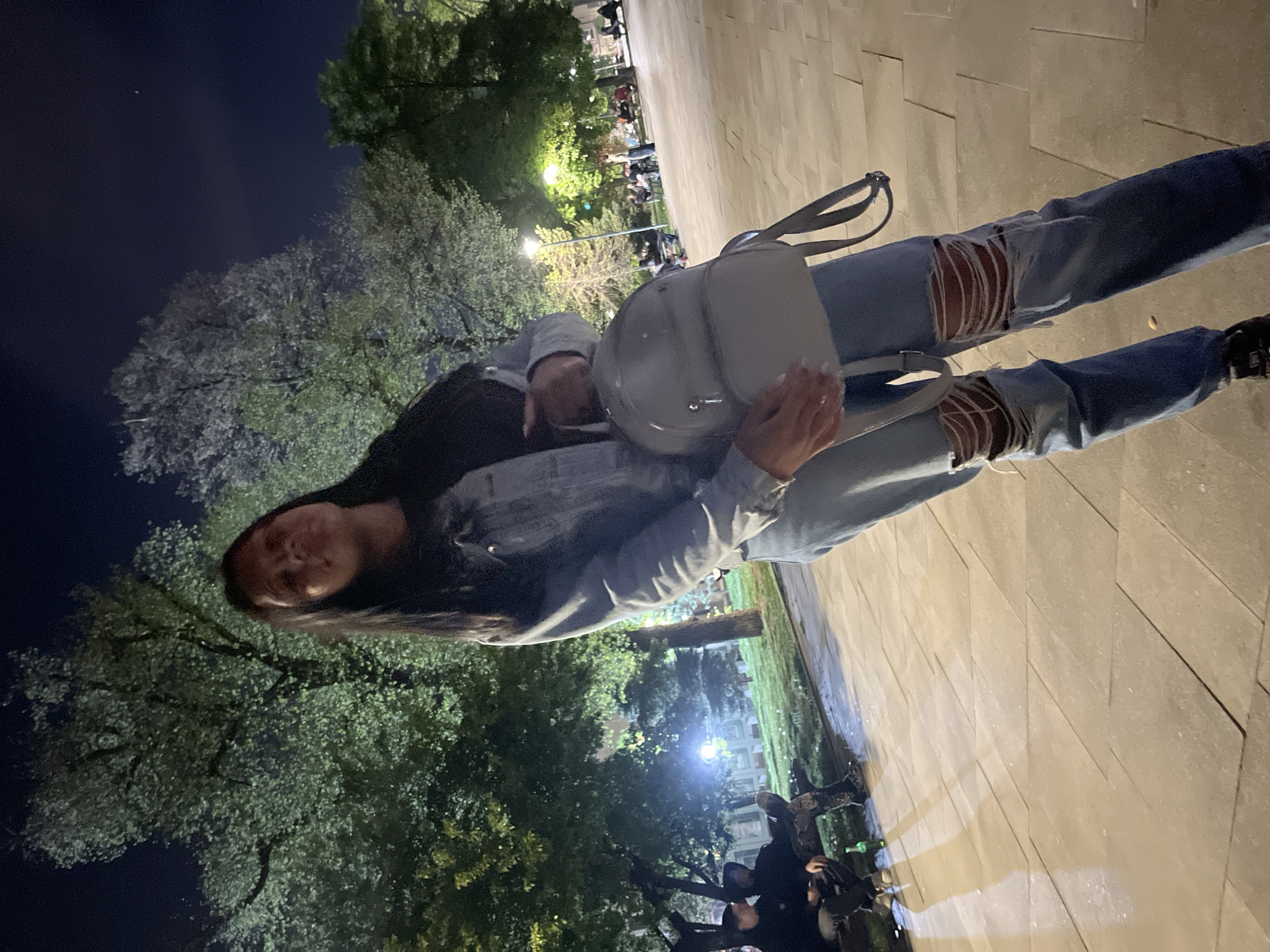 The media file features a full-length portrait of a young woman standing outdoors at night, likely in a park or plaza in Sofia, Bulgaria.
**Subjects:** The central subject is a woman with long, dark hair, looking directly at the camera with a slight smile. She is dressed in a black top, a light blue denim jacket, and light blue ripped jeans. She wears dark shoes and carries a light-colored (possibly white or light grey) shoulder bag. In the background, several other people are visible, mostly seated or standing in groups, appearing to be socializing.
**Scene and Setting:** The setting is an urban outdoor space, characterized by a paved ground made of light-toned rectangular tiles. Numerous trees with green foliage are present, suggesting a temperate season. The scene is illuminated by artificial streetlights, casting light on the trees and the ground, creating areas of shadow and light. The sky is dark, indicating nighttime, with a faint bright dot visible in one crop, possibly a star or a distant light source.
**Activity:** The woman is posing for the camera, standing still. The background figures appear to be engaged in casual social interaction or relaxation.
**Notable Details:** The time of day is clearly night. The ripped detailing on the woman's jeans is a prominent feature of her attire. The overall mood appears relaxed and social. There is no visible text in the image.