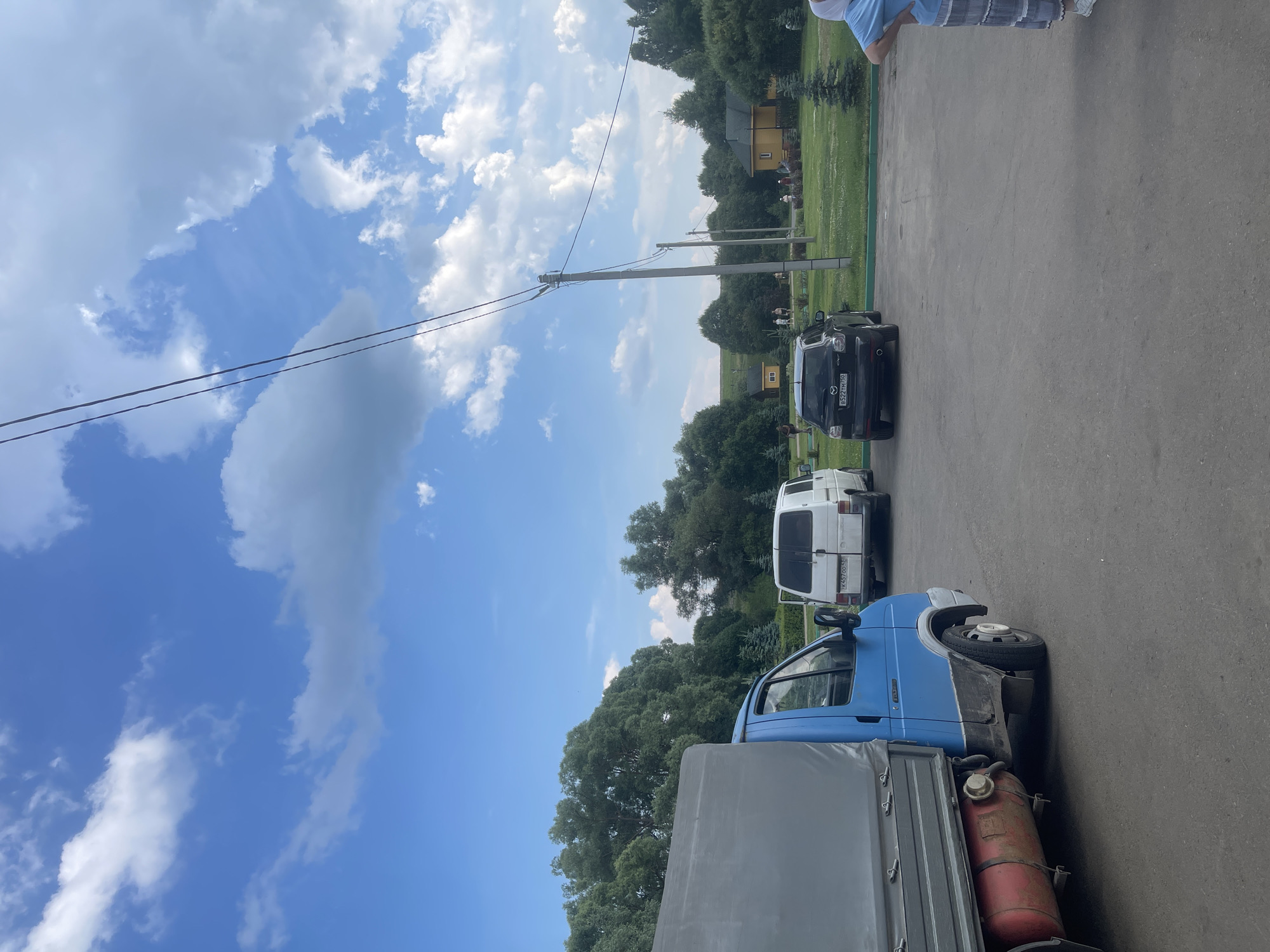 In the city of Кременки, Russia, a blue truck with a covered cargo bed is parked on asphalt. To its right, a white van is parked, and further to the right, a dark-colored SUV is present. In the background, there are green trees, a grassy area, two small yellow buildings, and a larger yellow building. Several people are visible in the background, walking in the grassy area. The sky above is blue with scattered white clouds. Power lines run across the upper portion of the image.