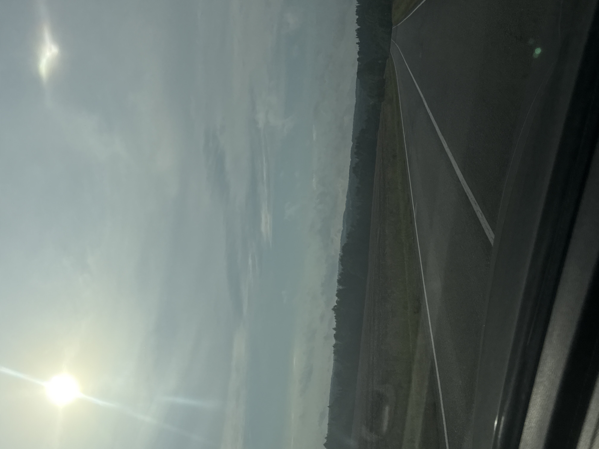 The image depicts a rural landscape with a road stretching into the distance, flanked by fields and a treeline. The sky is overcast with clouds, and the sun is visible as a bright, slightly diffused orb in the upper left corner, casting a lens flare. Another fainter, circular light effect, possibly a secondary lens flare or reflection, is present in the upper right. The road is paved with white lines marking its lanes. The foreground shows the dark interior of a vehicle, suggesting the image was taken from a car. The overall impression is one of travel through a serene, natural environment. The location is identified as Nylga, Russia.