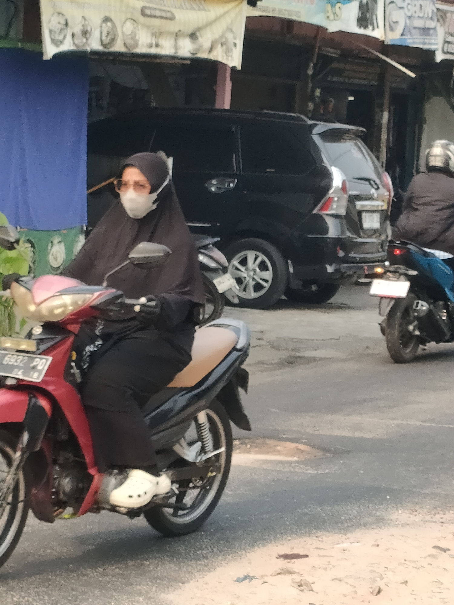 A woman wearing a black hijab and a white face mask rides a red motorcycle. She is wearing a black top, black pants, and white Crocs-style shoes. The motorcycle has a license plate that reads "6932 PO". Behind her, a black SUV is parked. The scene appears to be on a street in Pekanbaru, Indonesia, with buildings and shops visible in the background. The ground is paved, with some debris and patches of dirt. A second person on a blue motorcycle is visible in the far right of the image.
