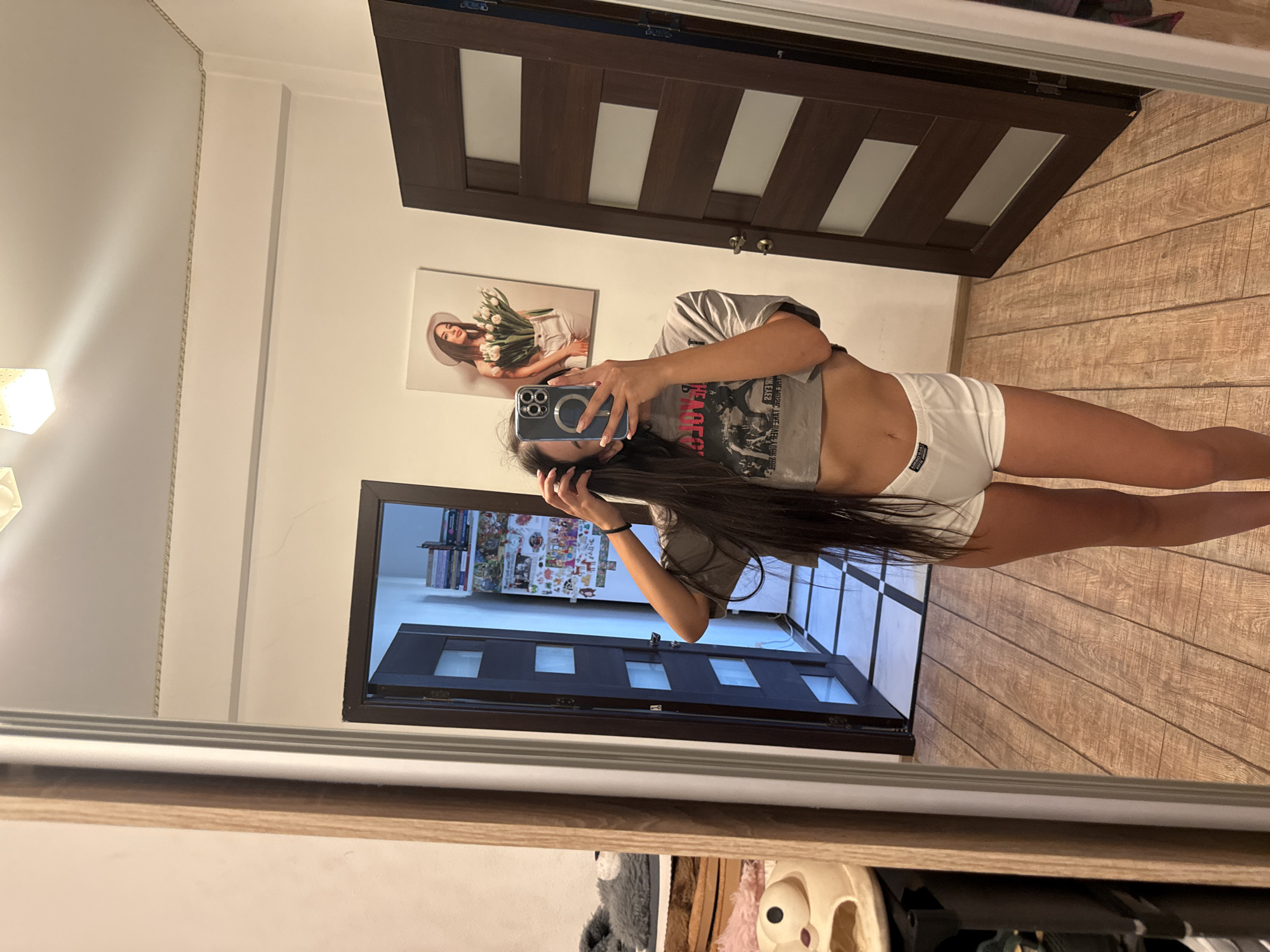 The image shows a young woman taking a selfie in a mirror, as reflected in the mirror. She is wearing a grey t-shirt and white shorts. The background includes a wooden door, a painting of a woman with flowers, and a bookshelf. The scene appears to be indoors, likely in a home. The lighting suggests it could be daytime. The woman's expression is neutral as she focuses on taking the photo. The image has a casual and personal feel. There is text on the woman's t-shirt, but it is reversed and difficult to read. The overall setting is a residential interior.