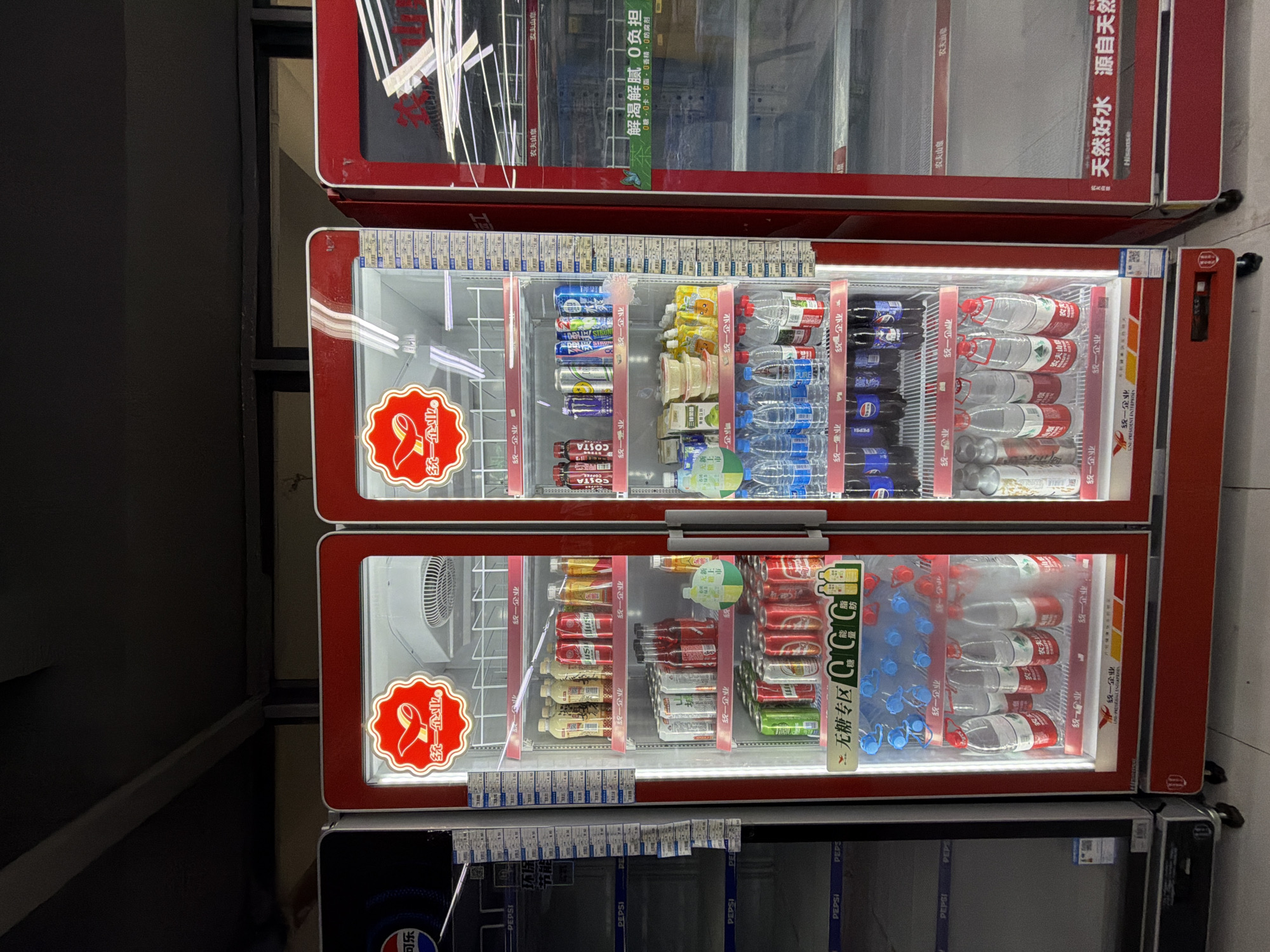 The image shows a row of commercial refrigerators in a retail setting, likely a convenience store or supermarket, in Jiangjin, China. The refrigerators are primarily red with clear glass doors displaying a variety of beverages and some food items.
The refrigerator on the far left is partially visible and appears to be a Pepsi branded cooler, with several shelves containing Pepsi products. The middle and right refrigerators are open and fully visible.
The middle refrigerator is a double-doored unit. The top shelves on the left side display bottles of what appear to be tea or juice in yellow and red packaging, alongside cans of what look like soda. Further down, there are rows of canned beverages, including what appear to be beer and soda cans in red, silver, and green. The lower shelves on the left contain bottles of water. The top shelves on the right side of the middle refrigerator display cans of Costa coffee and other beverages. Below these are small bowls of food items, likely snacks or desserts, followed by bottles of water.
The rightmost refrigerator is also a double-doored unit, but its shelves are mostly empty, with only a few items visible on the top shelf. A prominent sign on the lower part of this refrigerator reads "天然好水 源自天然" which translates to "Natural good water, from nature."
Decorative logos featuring a red bird-like emblem with the text "统一企业" (Uni-President Enterprises) are visible on the upper portion of the middle and right refrigerators. Additionally, there are price tags and product information labels displayed on the shelves and doors of the refrigerators. The overall environment appears to be indoors, with overhead lighting visible. The floor beneath the refrigerators is tiled. The refrigerators are equipped with wheels for mobility.