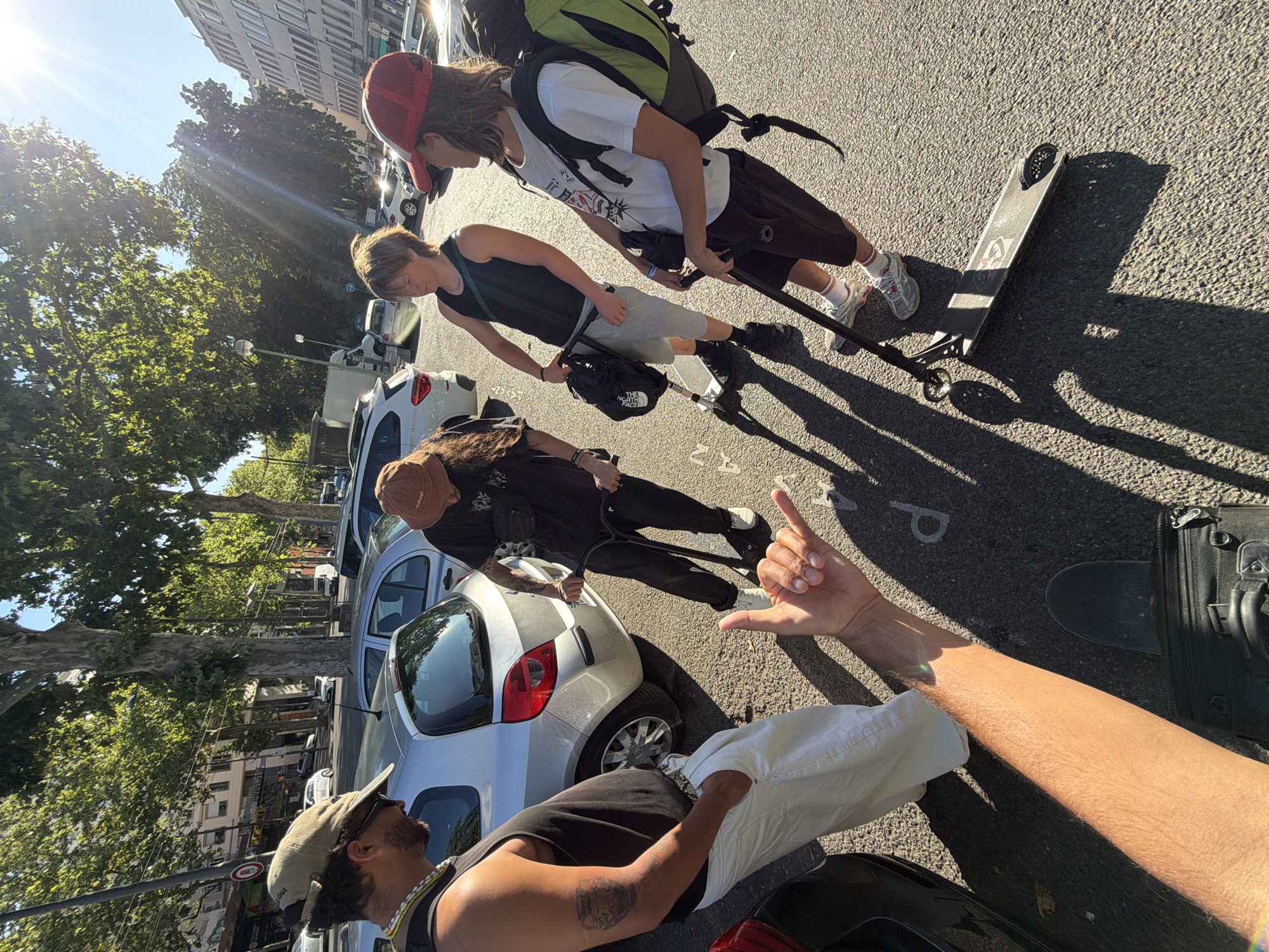 A group of four young adults, two males and two females, are standing on an asphalt street in a city. The person in the foreground, on the left, is a male wearing a black tank top, white pants, and a tan baseball cap. He is looking down and has his right hand in his pocket. In front of him, a male is performing a "shaka" hand gesture. To his right, a male wearing a black tank top and grey shorts is holding onto a black scooter. To the right of that, a male wearing a white t-shirt and brown shorts, with a large green and black backpack, is also holding a scooter. Another male, wearing a brown bucket hat and a black t-shirt, is on a black scooter to the left of the tank-top wearing male. The background shows cars parked along the street, trees with green foliage, and buildings. The scene appears to be during the daytime, with sunlight casting shadows on the ground. The presence of trees, architecture, and vehicles suggests an urban environment. Specifically, this scene depicts a street in the city of Lyon, France, with the architecture and streetscape characteristic of the area.