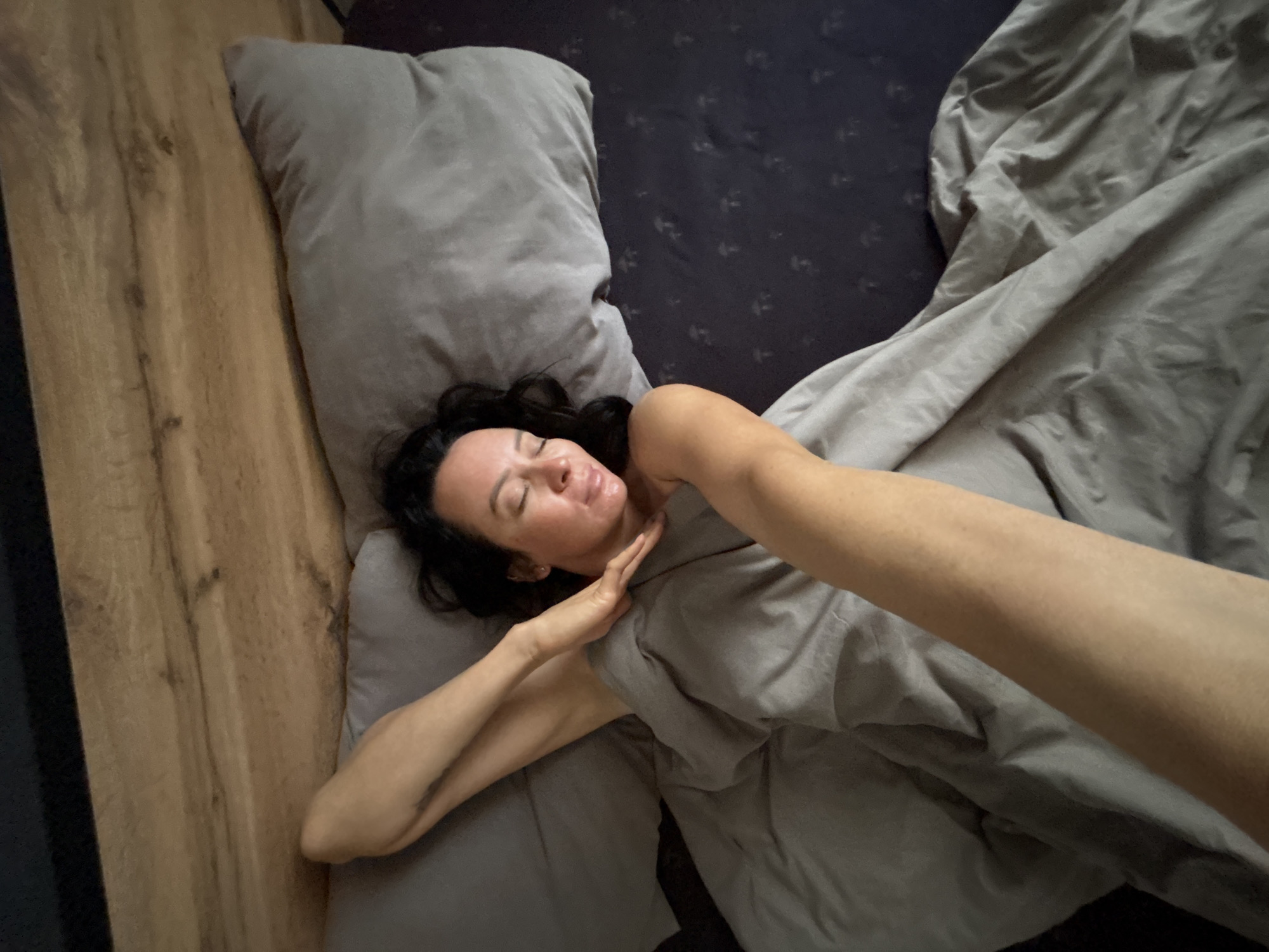 A woman with dark hair lies on a bed in an interior setting. She rests on a gray pillow atop a dark patterned bedsheet. Part of a wooden floor is visible to the left of the pillow. Gray bedding covers the lower portion of her body. Her left arm rests across her chest and neck, and her right arm extends into the foreground. The setting is possibly a private residence in a city such as Chaikovsky, Russia, but there is no explicit evidence of the city in the image.