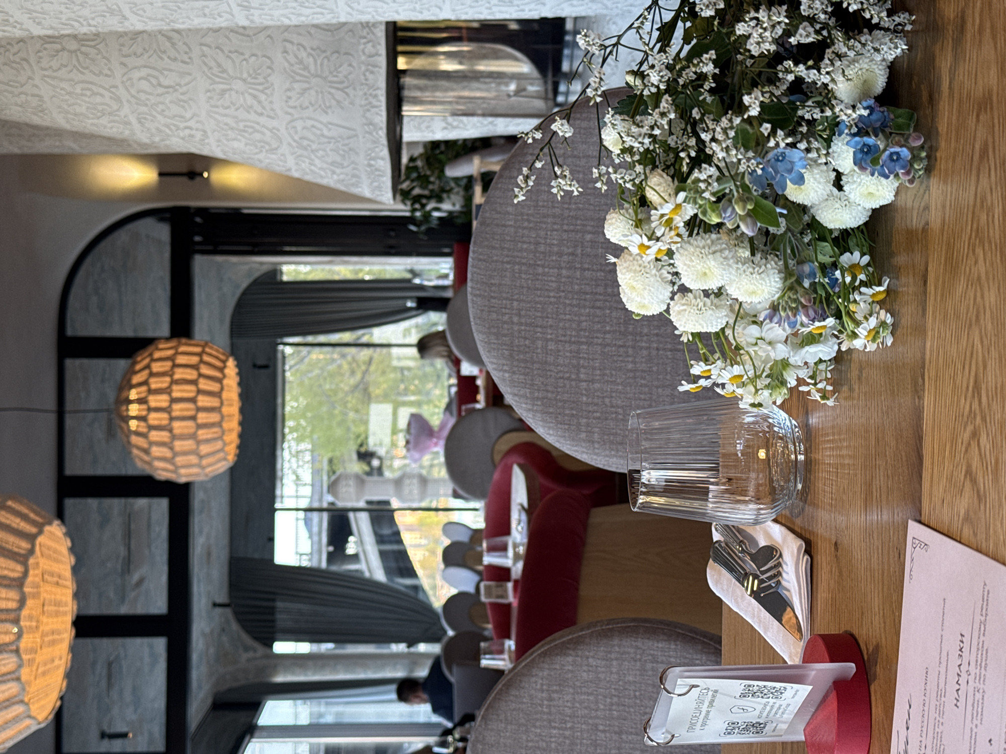 A slightly overhead, medium shot shows a table arrangement inside a restaurant, facing outward toward a window and people, in Krasnoyarsk, Russia. A grey, textured chair dominates the left side of the frame. In the center, a water glass, silverware on a napkin, and a menu on a red base sits on the wooden table. A bouquet of white flowers sits between the chair and the objects on the table. In the background, a row of upholstered chairs, a table, and lighting. A large window looks outside to pedestrians on the street. An arched ceiling hangs above.