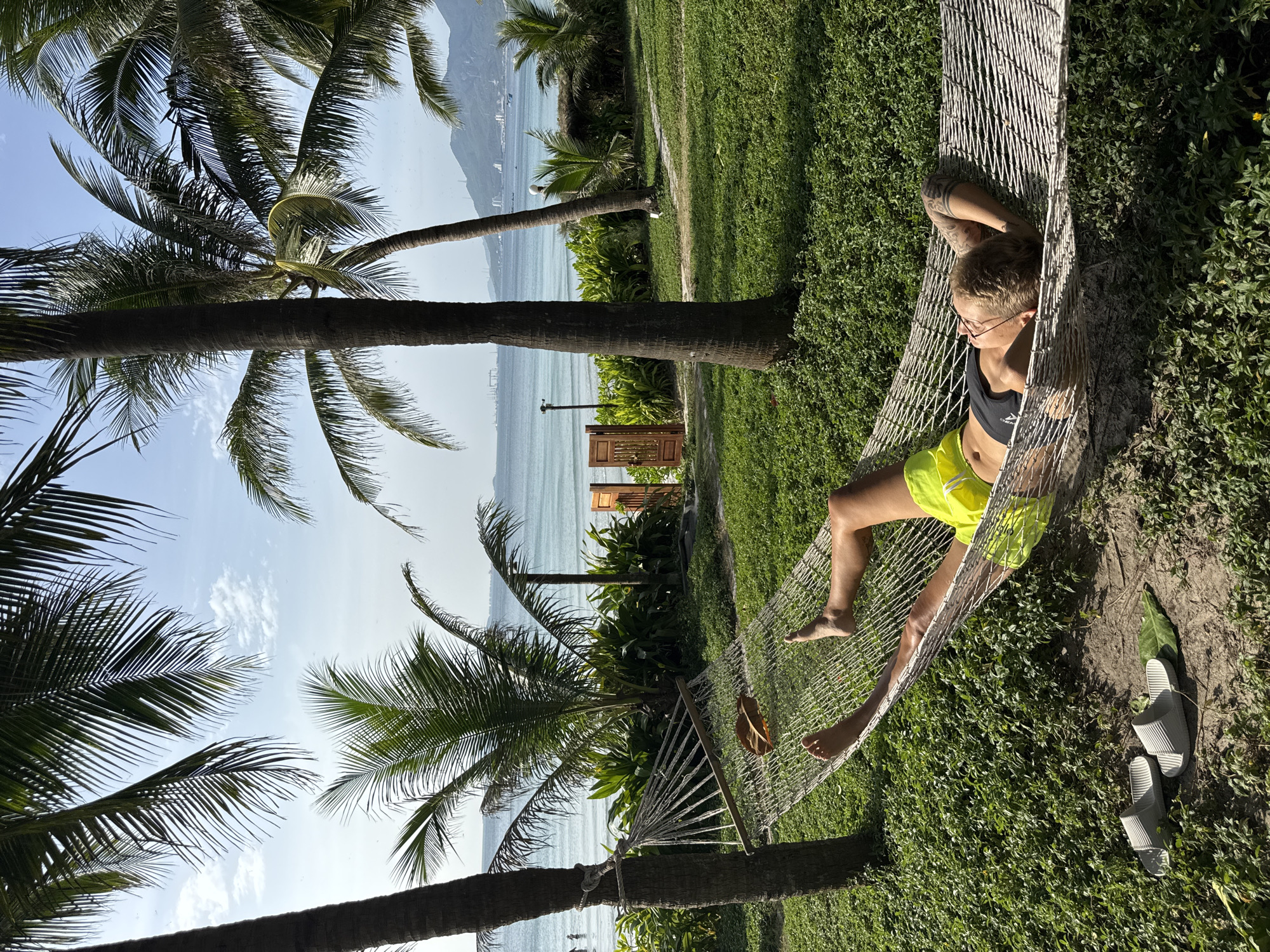 A person is relaxing in a hammock strung between two palm trees on a grassy area overlooking the ocean. The person is wearing a black tank top and neon yellow shorts. Their head is resting on their arms, and their eyes are closed. Two pairs of white slides and a green leaf are on the ground near the hammock. In the background, the ocean is visible with a few boats on the horizon. There are lush green plants and more palm trees surrounding the hammock and leading down to the beach. The sky is blue with a few scattered clouds. The location is identified as Đông Ninh Hòa, Vietnam.