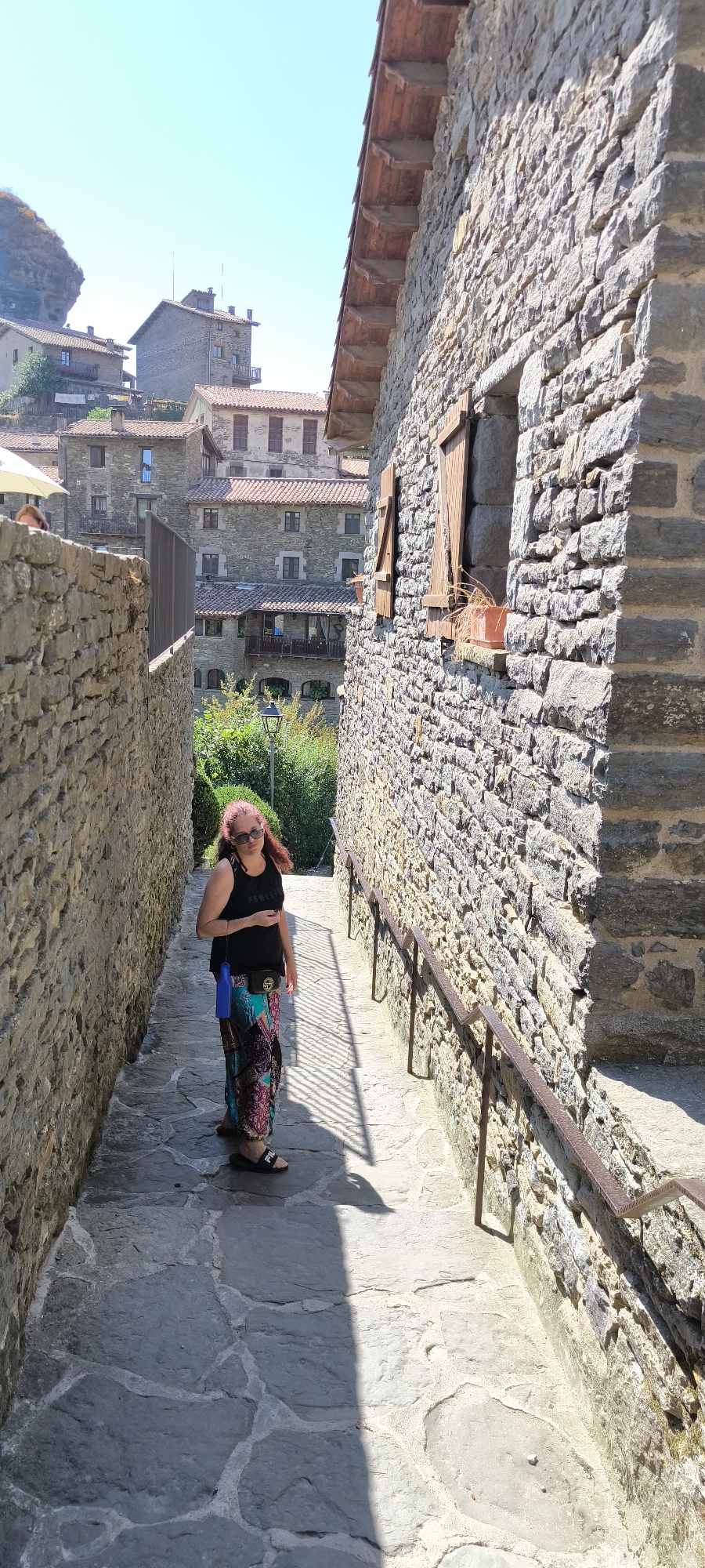 A woman with reddish-brown hair and sunglasses stands in a narrow cobblestone alleyway in Rupit i Pruit, Spain. She is wearing a black tank top, a colorful patterned skirt, and black flip-flops. Around her waist, she has a small black fanny pack. In her right hand, she holds a smartphone. The alleyway is flanked by stone buildings with wooden shutters. In the background, other stone buildings with red tile roofs are visible, characteristic of the medieval village. The sky is clear and blue, indicating a sunny day. A metal handrail runs along the right side of the alley.