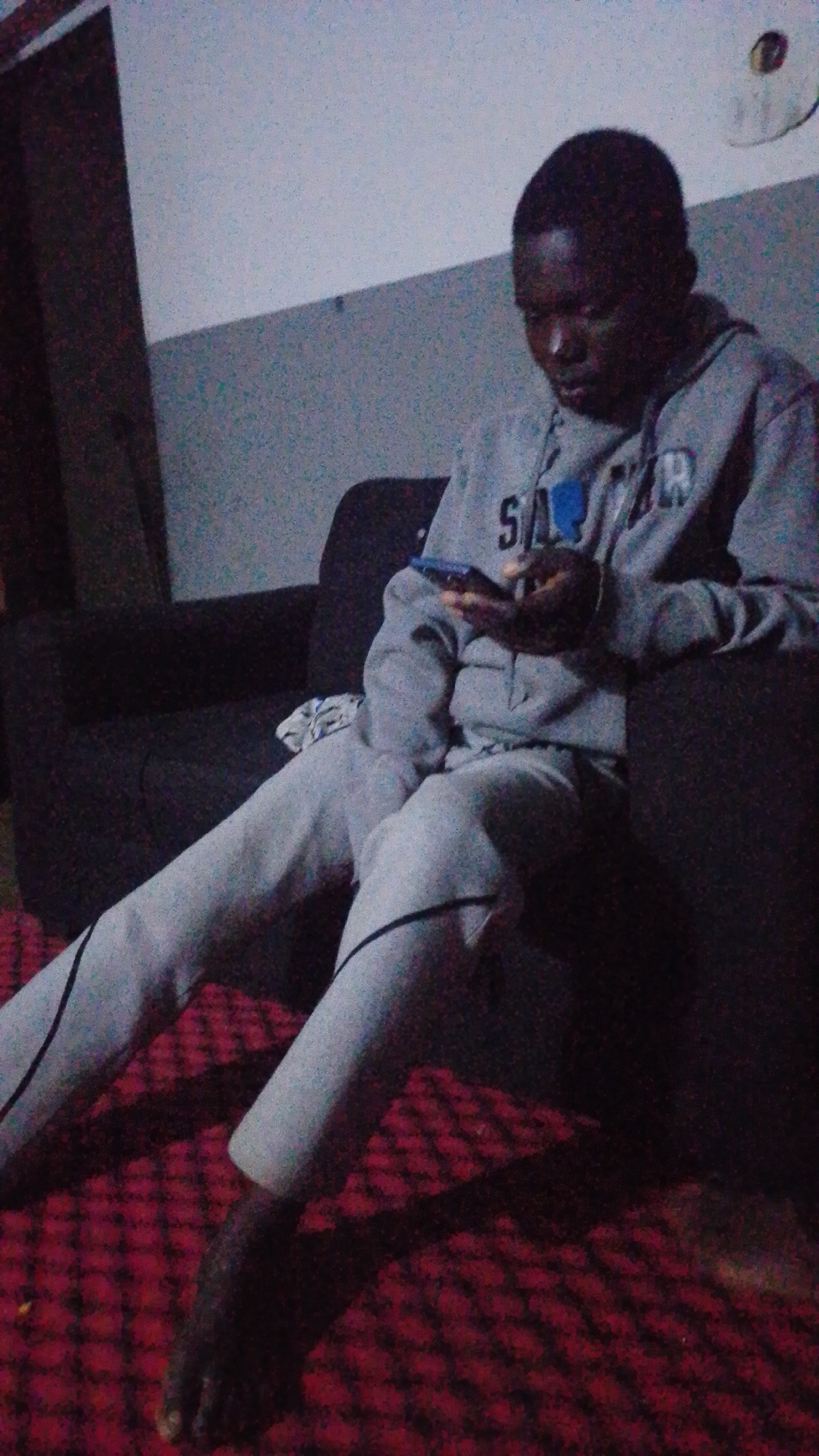 The image shows a young Black male, likely a teenager, sitting on a dark-colored couch with his legs extended and crossed. He is wearing a light grey hooded sweatshirt with the word "SA" visible on the chest in black lettering, and light grey sweatpants with black stripes down the sides. He is holding a dark-colored smartphone in his right hand, looking down at it intently. The background consists of a light-colored wall with a darker, horizontal band across it. A circular object, possibly a clock or a decorative item, is mounted high on the wall to the right. The foreground is dominated by a red, textured rug with a diamond pattern. The lighting in the room appears to be dim, creating shadows. The scene is consistent with an indoor residential setting in Jos, Nigeria, where such furniture, clothing, and electronic devices are common.