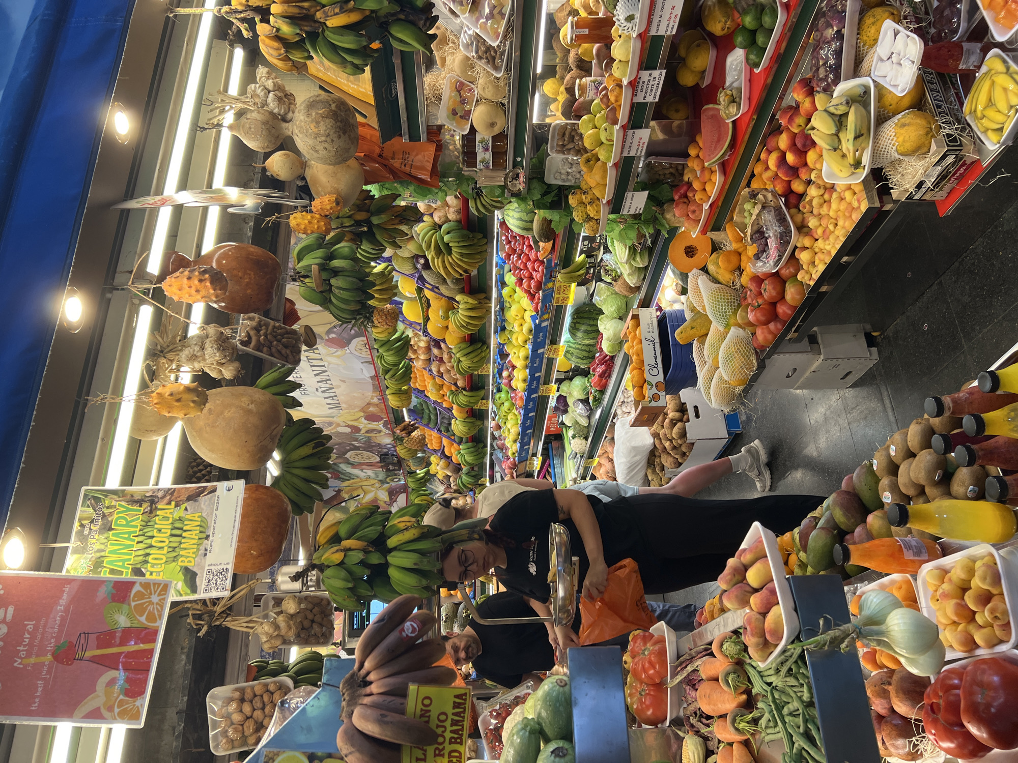 Here is a description of the image:
The image shows a vibrant, bustling fruit and vegetable market in Las Palmas, Spain. The scene is filled with a colorful array of fruits and vegetables, meticulously arranged in stalls. Bananas, melons, tomatoes, peaches, and other produce are abundantly displayed.
People are present, including a woman in a black shirt wearing glasses, who appears to be weighing produce. Another individual, also wearing a black shirt, is nearby. There are other people in the background, including one person wearing light colored shorts and white sneakers.
Signs with descriptions and prices are visible throughout the market, including "CANARY ECOLOGICAL BANANA" and "ROJO RED BANANA." The image has a slight tilt, giving the impression of looking upward or downward along the length of the market stall. Artificial lights are visible on the ceiling of the marketplace.