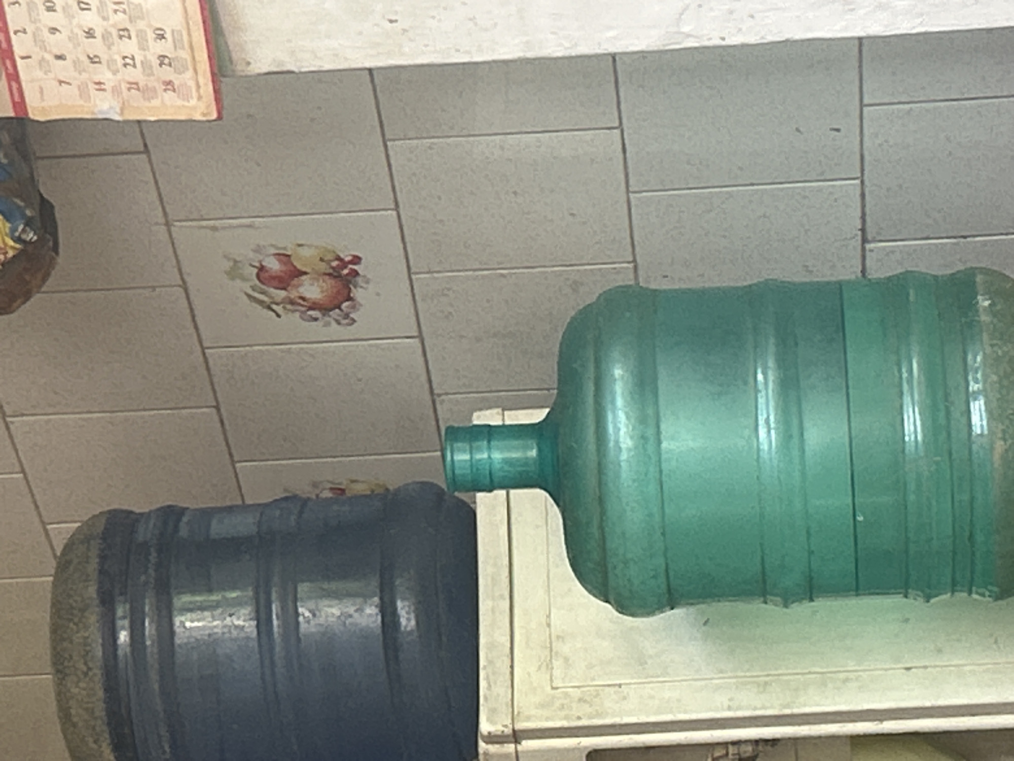 Two large water jugs are visible in this image. The one in the foreground is teal colored and appears to be made of plastic, with a narrow neck. It sits on top of a white cabinet. Behind and to the left of the teal jug is a dark blue water jug, also large and likely made of plastic, with a textured lid. The jugs are placed against a tiled wall. A calendar with visible dates from 1 to 31 is hanging on the wall to the right. A small decorative tile with an image of fruit, possibly apples and grapes, is also affixed to the wall. The overall environment suggests an indoor domestic or possibly commercial space. There are no people visible in the image. The location is identified as Santa Ana, Venezuela.