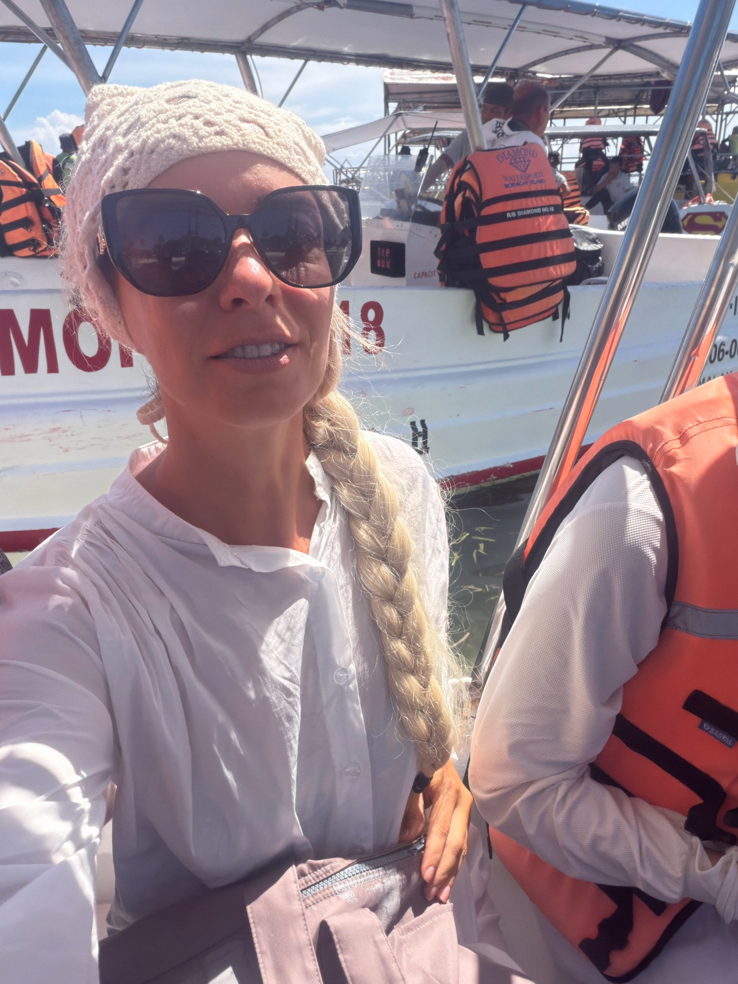 In Malay, Philippines, a woman with blonde hair in a braid wears a white crocheted hat and large black sunglasses. She is on a boat, as indicated by the railing and the white side of the boat with red lettering that reads "MON..." and "06-08". She is wearing a loose white shirt and a pink bag is visible. Another person in an orange and black life vest is seated next to her on the right. In the background, other boats and people wearing life vests are visible, suggesting a tourist activity in a coastal area. The lighting indicates it is daytime.