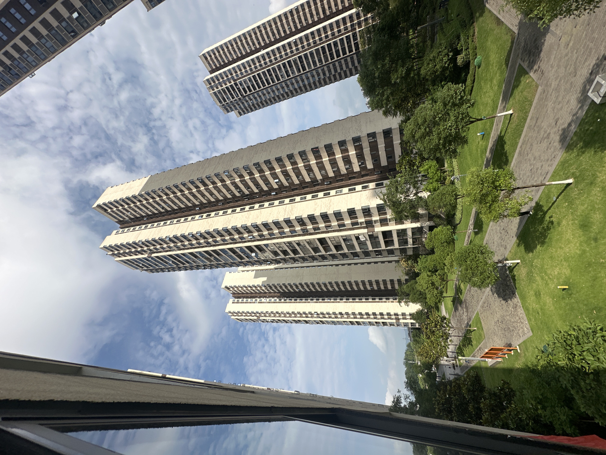 The image shows a view from a high-rise building in the Chongqing High-tech Zone, China. Several other high-rise residential buildings are visible, interspersed with green spaces consisting of trees and lawns. A pedestrian walkway zigzags through the green areas. The sky is partly cloudy. The view is from inside a building, as evidenced by a window frame and its reflection in the lower portion of the image.
