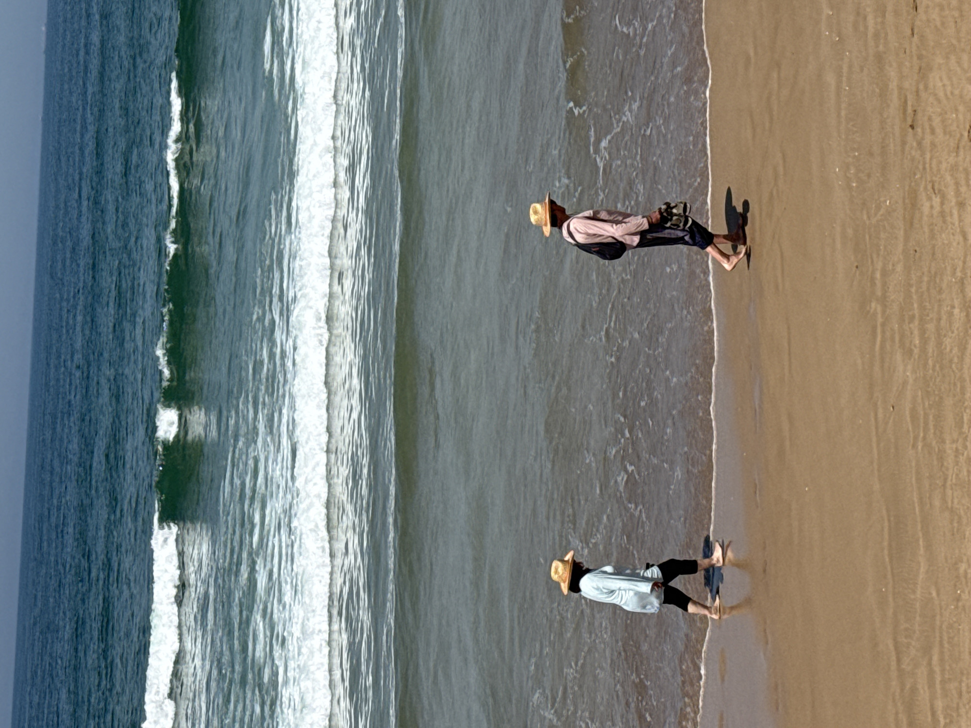 Two individuals are walking barefoot on a sandy beach alongside the ocean in Agadir, Morocco. Both are wearing hats. One individual is wearing a light-colored long-sleeved shirt, dark pants, and walking near the edge of the surf. The other individual is wearing a light-colored shirt, dark pants, and carrying a dark backpack. Waves are rolling onto the shore. The sky is overcast.