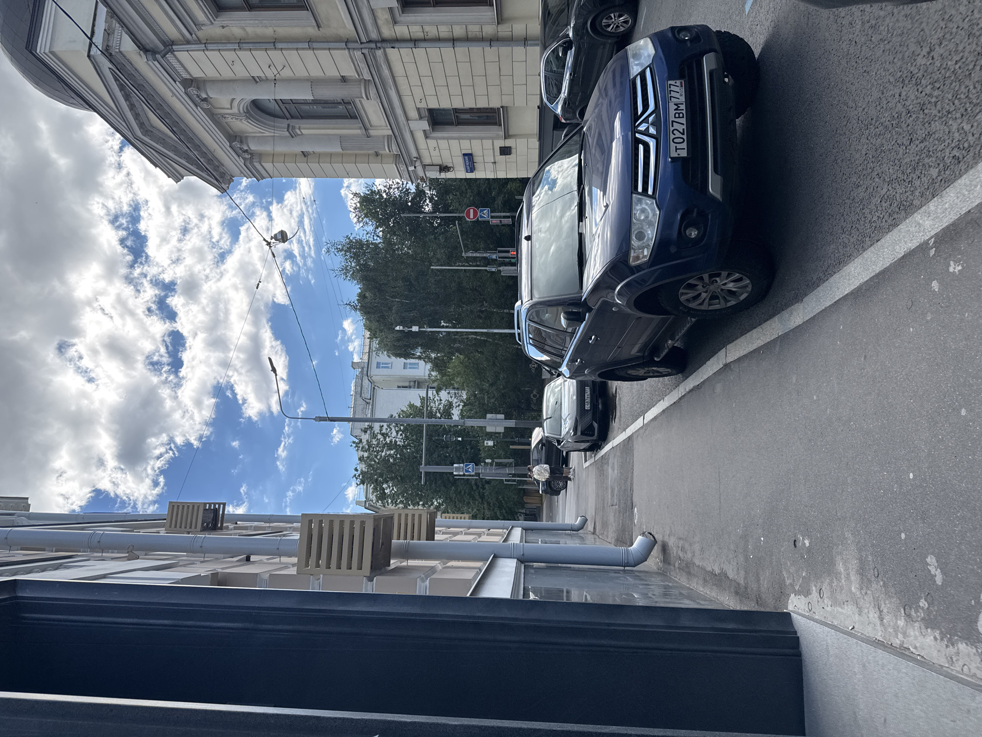 A blue Mitsubishi Pajero is parked on a city street in Moscow, Russia. The vehicle's license plate is visible and reads "T027BM777". The Pajero is in the foreground, facing right, with its front end slightly angled towards the camera. To the left of the Pajero, a dark gray car is parked, and further down the street, another vehicle is visible. A pedestrian is walking on the sidewalk to the left of the parked cars. The street is lined with buildings on the left and right sides. The building on the right has a classical architectural style with columns and arched windows. The sky is cloudy with patches of blue. The scene appears to be a typical urban environment.