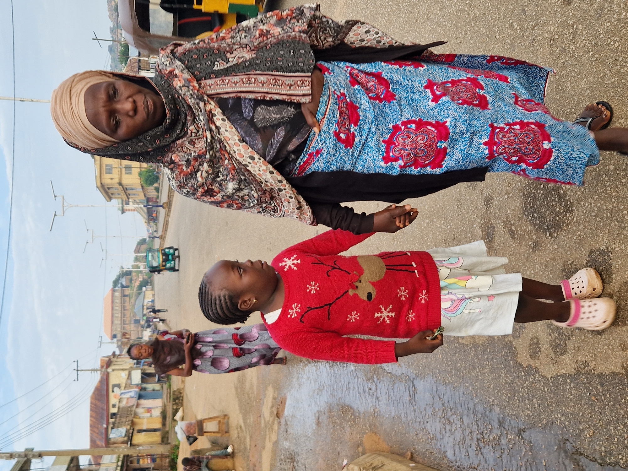 A woman and a young girl walk side-by-side on a wet street in Kabong, Nigeria. The woman, on the right, wears a light-colored headscarf and a patterned shawl over a black top and a blue and red patterned skirt. She holds the girl's hand. The girl, on the left, wears a red sweater with a reindeer and snowflake design, a white skirt with unicorn prints, and pink crocs. She is looking to her right. In the background, other people, buildings, and a green tricycle are visible along the street. The ground appears wet, possibly from recent rain.
