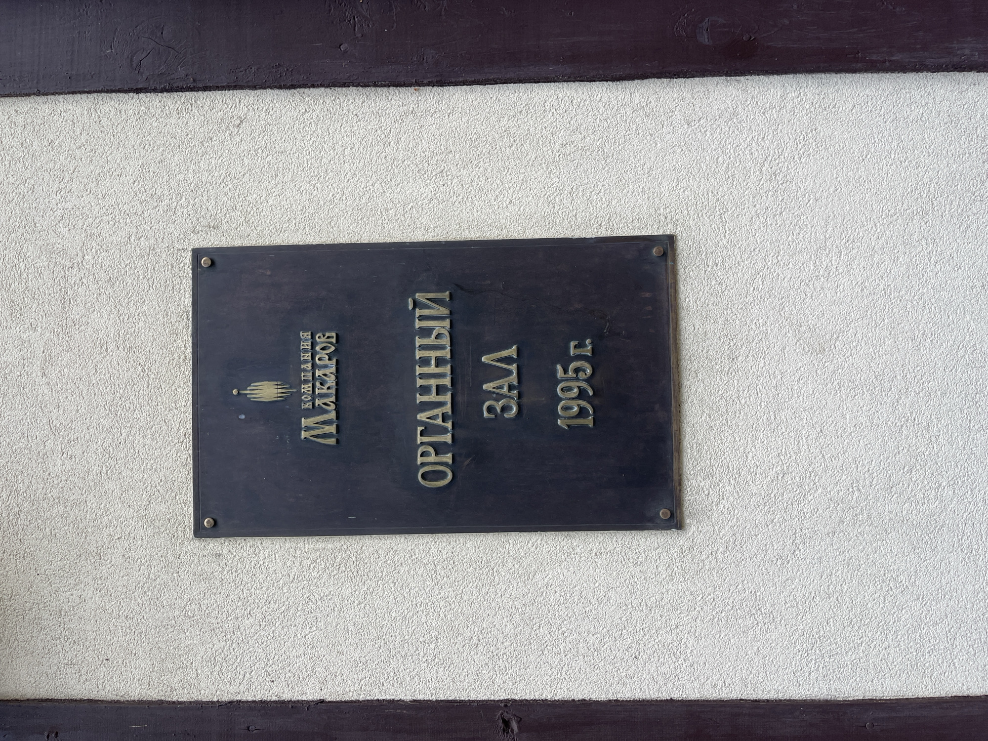 A dark rectangular plaque is mounted on a textured, off-white wall, possibly stucco, between two dark brown wooden posts. The plaque is inscribed with gold-colored lettering. At the top, a stylized gold emblem resembling a flame or a hand is centered above the word "МАКАРОВ" which is preceded by "КОМПАНИЯ" in smaller text. Below this, the phrase "ОРГАННЫЙ ЗАЛ" (Organ Hall) is written in large, bold letters. At the bottom of the plaque, the year "1995 г." is displayed. The plaque is secured to the wall by visible screws or rivets at its corners. The image appears to be taken in daylight. The location is the city of Svetlogorsk, Russia, as indicated by the plaque's inscription.