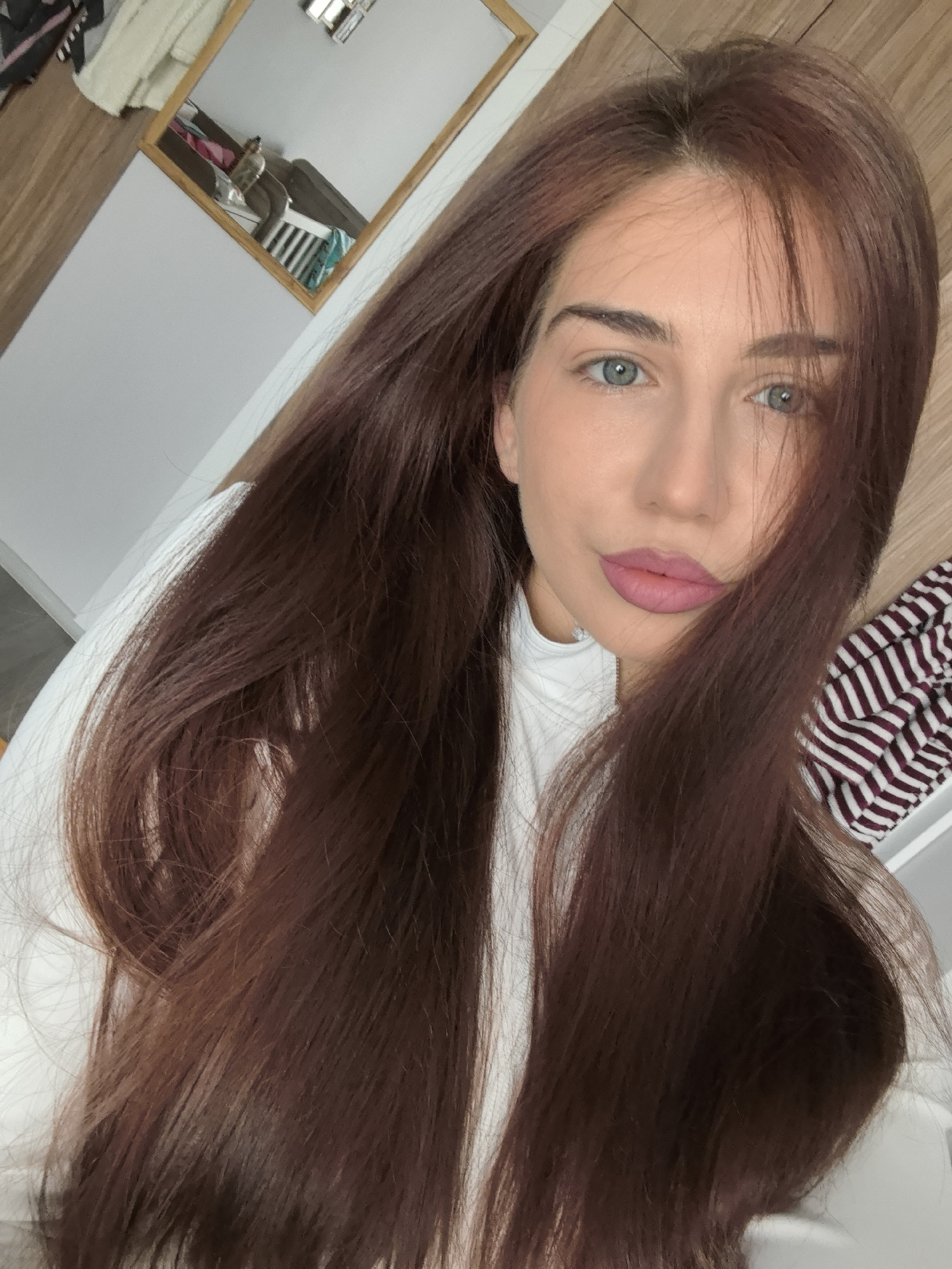 A young woman is captured in a close-up selfie, looking directly into the camera with a neutral to slightly serene expression. She has long, straight, dark brown hair that cascades over her shoulders, with some strands partially obscuring the right side of her face. Her striking blue eyes are a focal point, complemented by her full lips adorned with pink lipstick. She is wearing a white top, possibly a turtleneck.
The setting appears to be indoors, likely a personal space such as a bedroom in Stara Zagora, Bulgaria. In the upper left background, a wooden-framed mirror is visible, reflecting a portion of the room. The reflection shows a blurred figure, possibly a person, seated or hunched over on what looks like a bed with a white headboard. Other elements in the background include light-colored walls and wooden surfaces. To the right, a crumpled piece of fabric with dark stripes on a lighter background is partially visible. The overall lighting suggests an indoor setting, possibly during the day. No text is visible in the image.